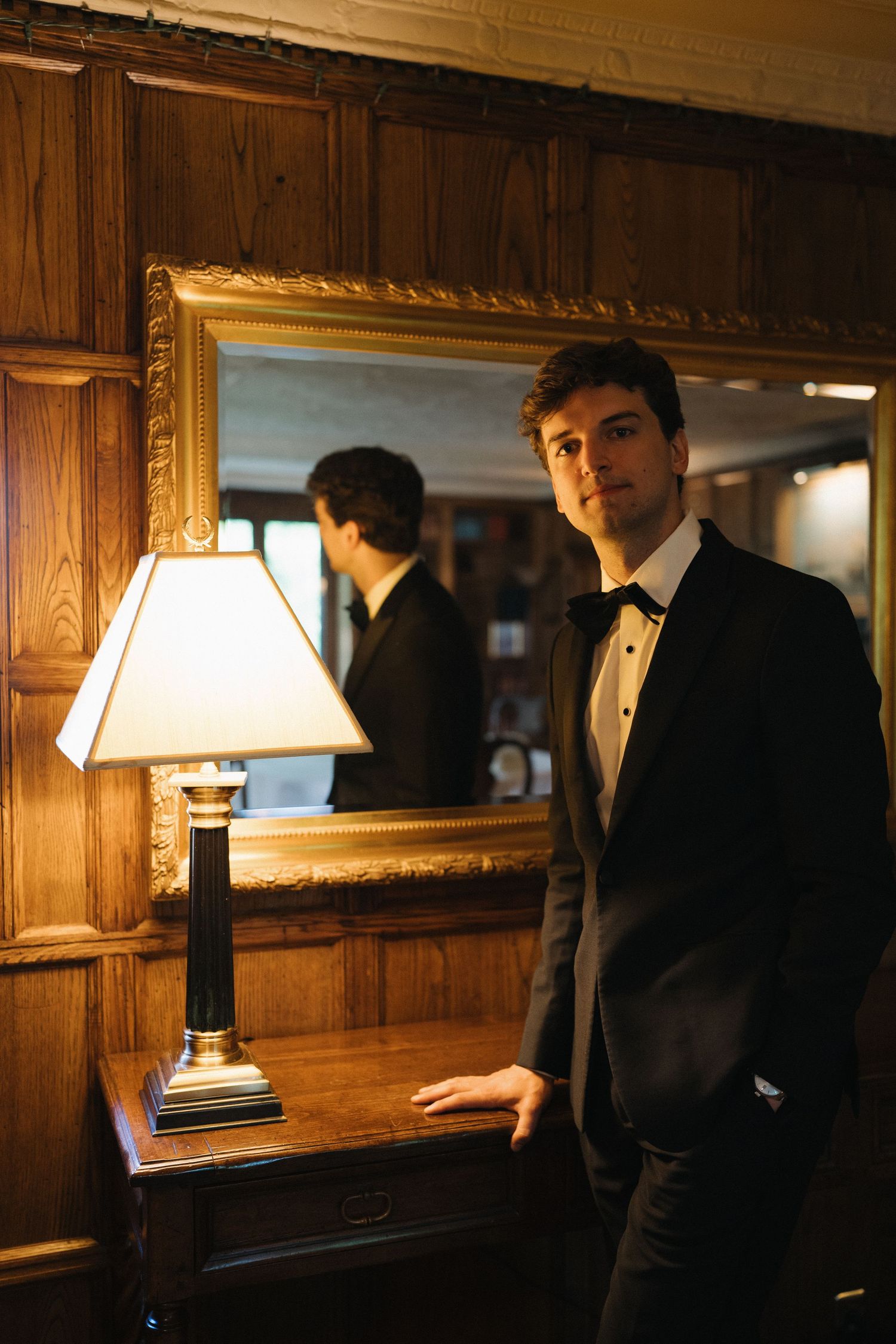 Groom in black tuxedo standing by ornate gold mirror and lamp in elegant wood-paneled room.