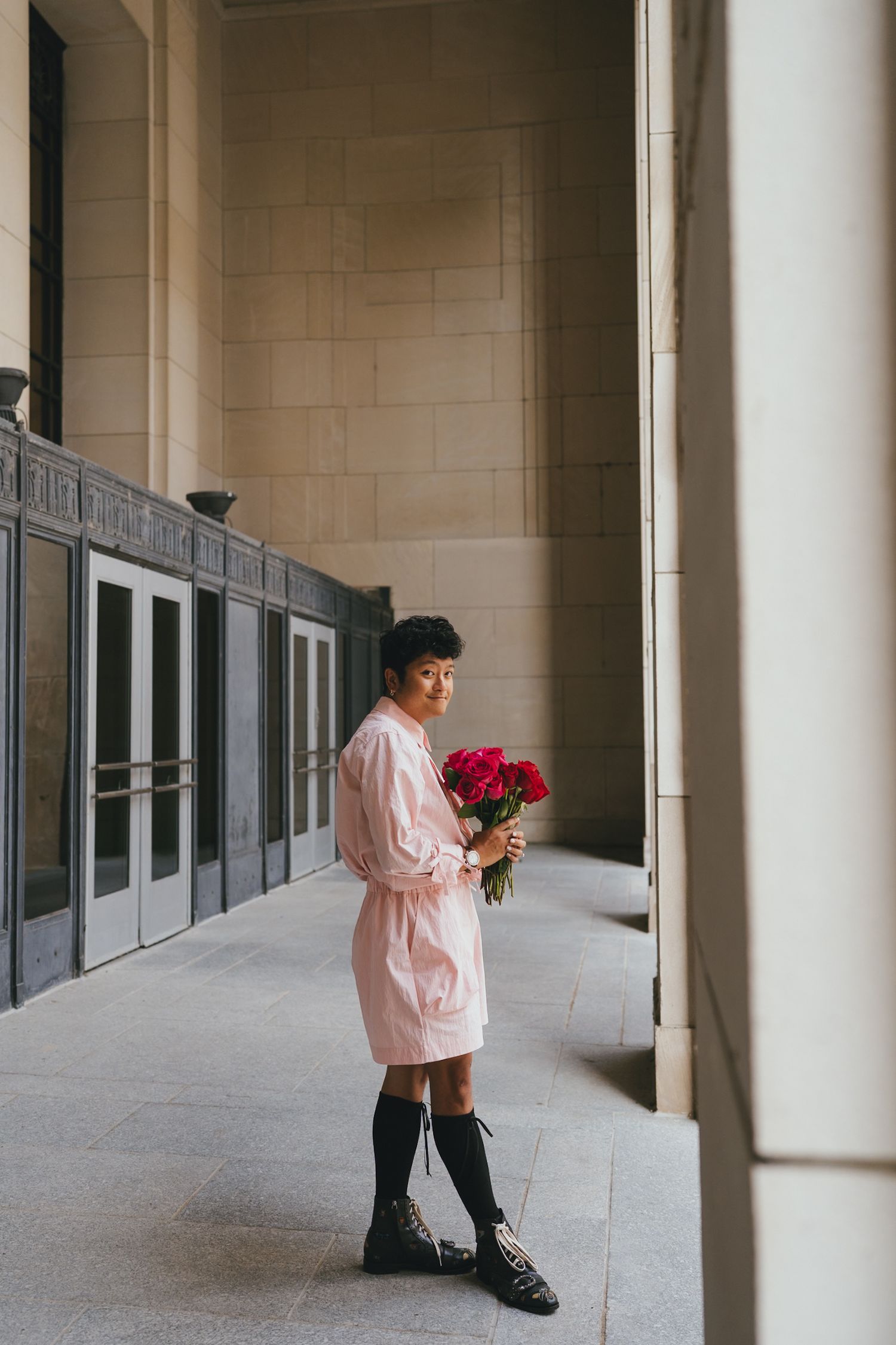 Woman in pink dress holding bouquet of red roses, wearing black knee-high socks and boots in elegant building corridor.