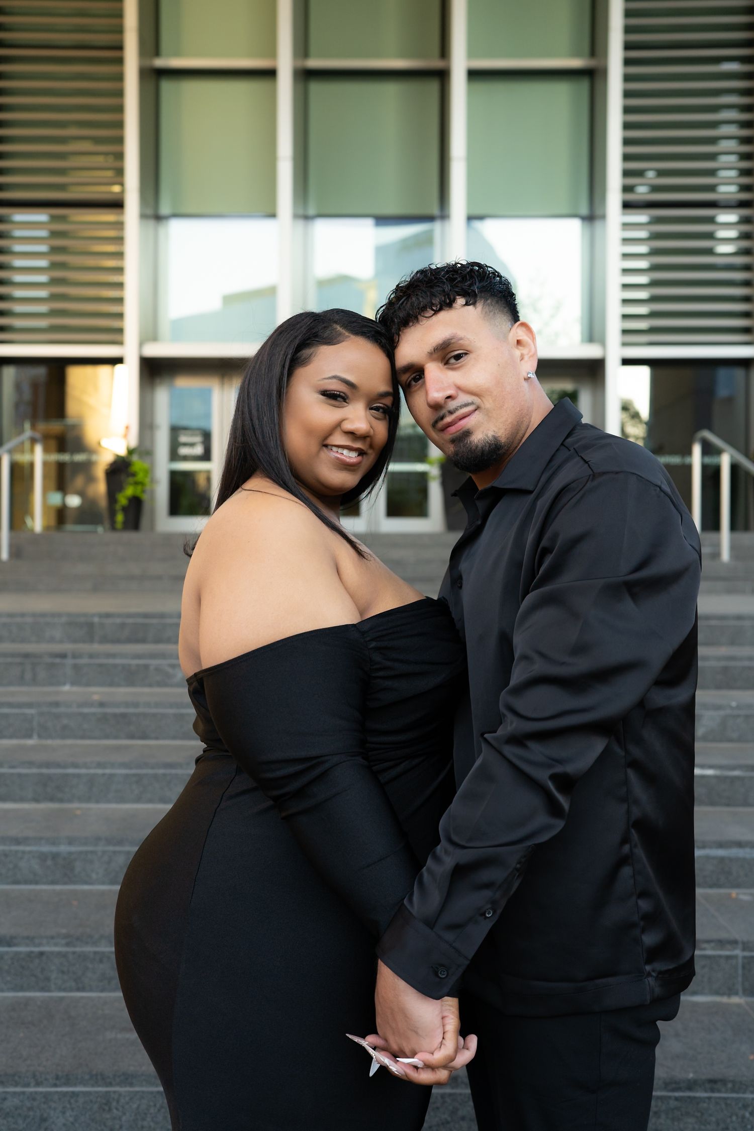 Smiling couple in black formal wear holding hands and posing in front of a modern building.