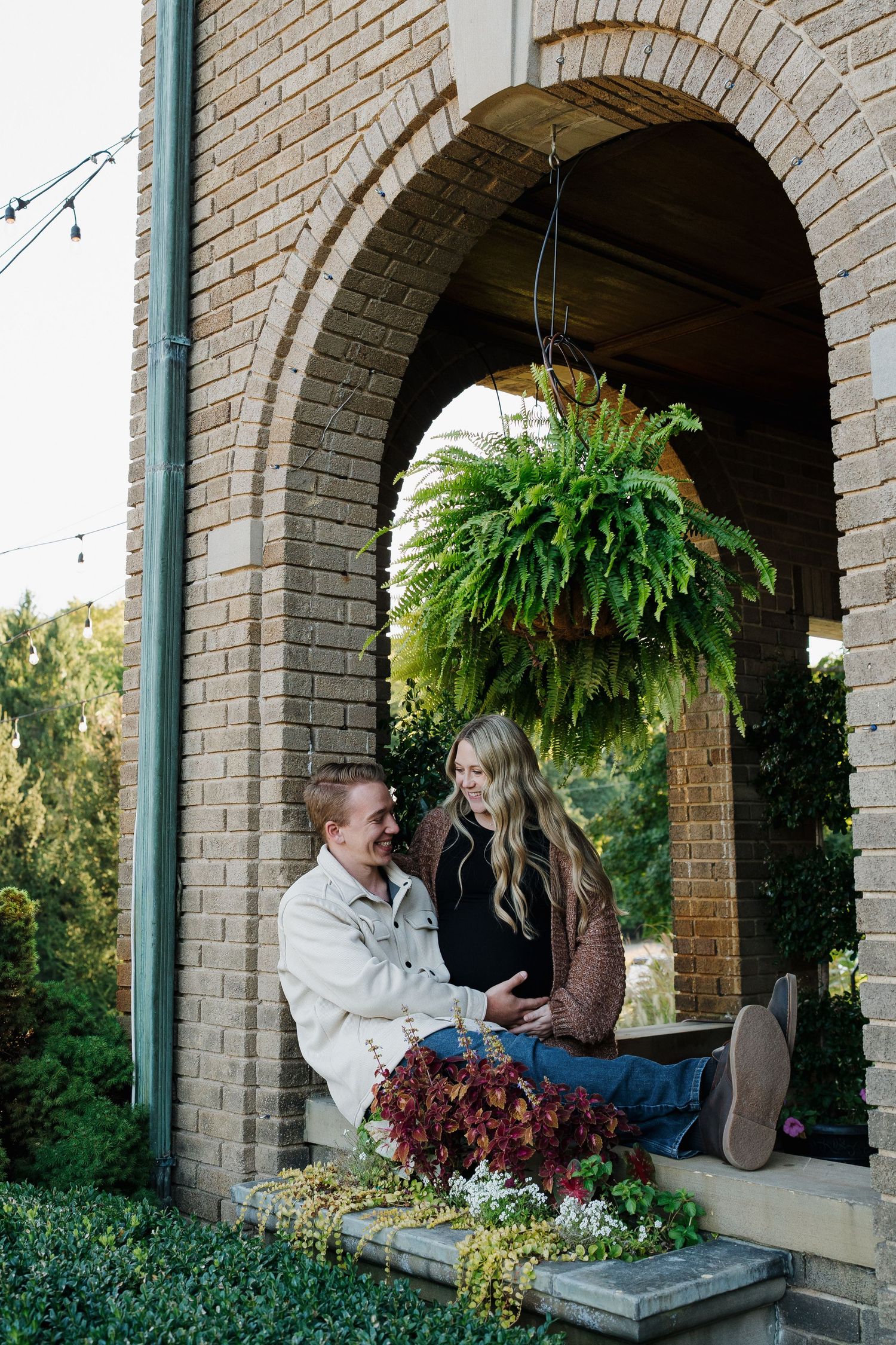 A smiling couple sits together under a brick arch with a hanging fern during maternity photos.