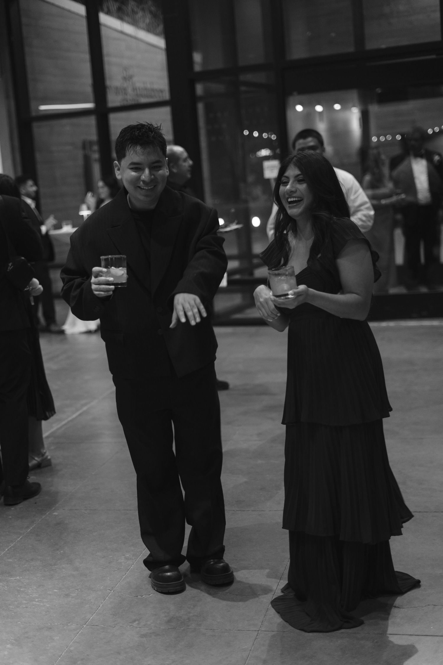 Laughing couple in black formal wear dancing and holding drinks at a lively wedding reception.