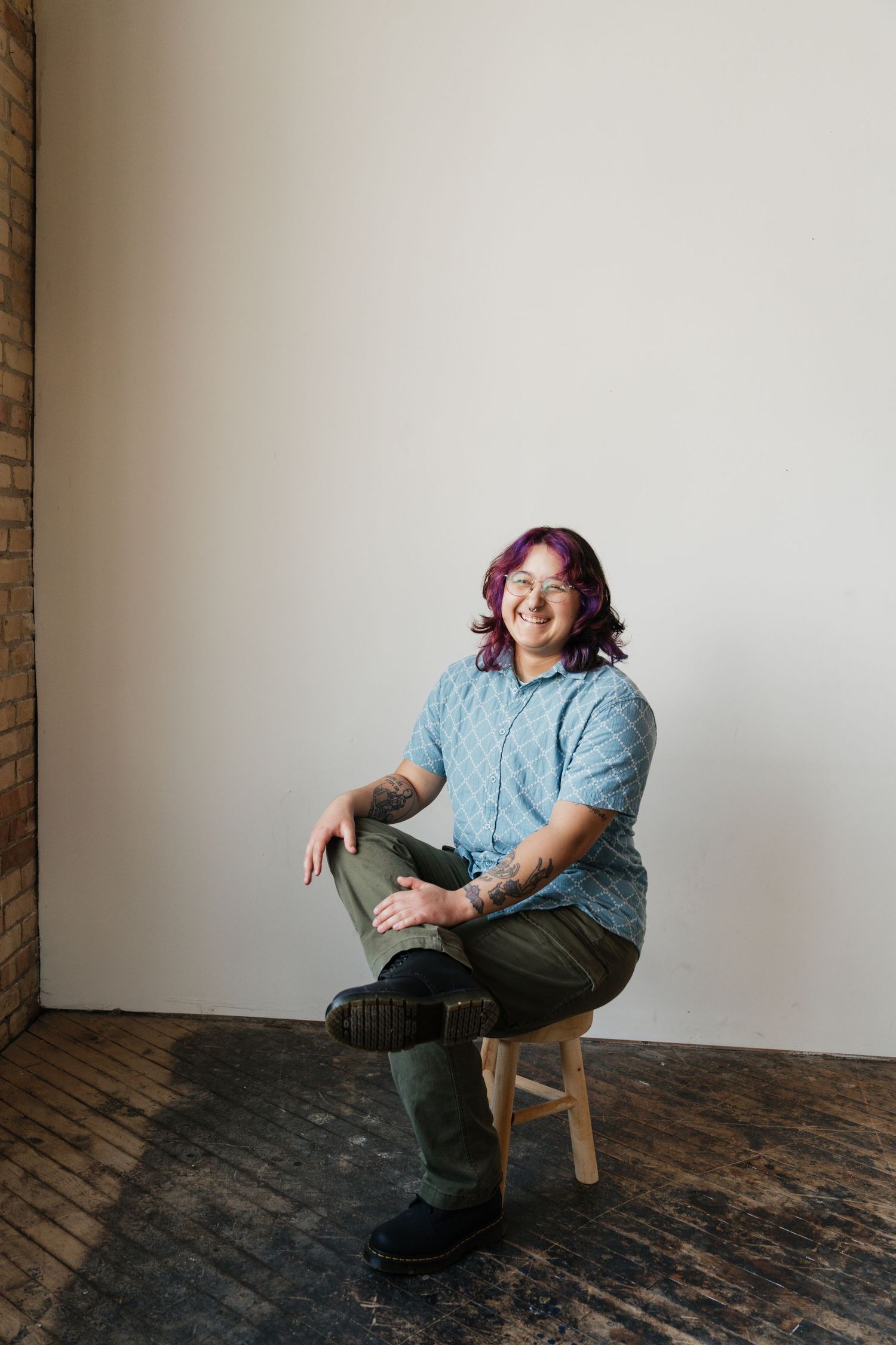 A person with purple hair and tattoos smiles while seated on a stool in a bright studio.