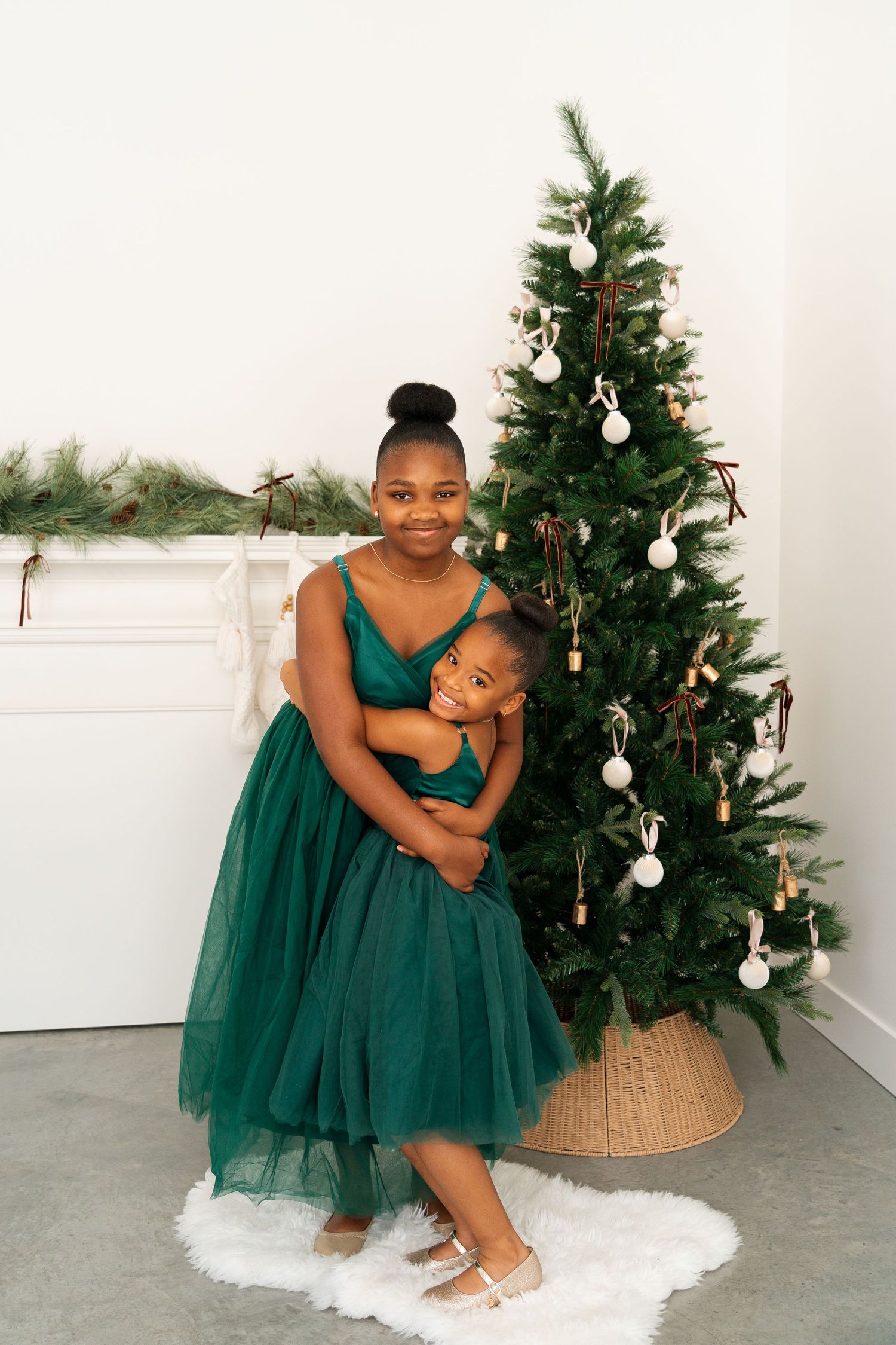 Two girls in matching green tulle dresses pose together beside a decorated Christmas tree.