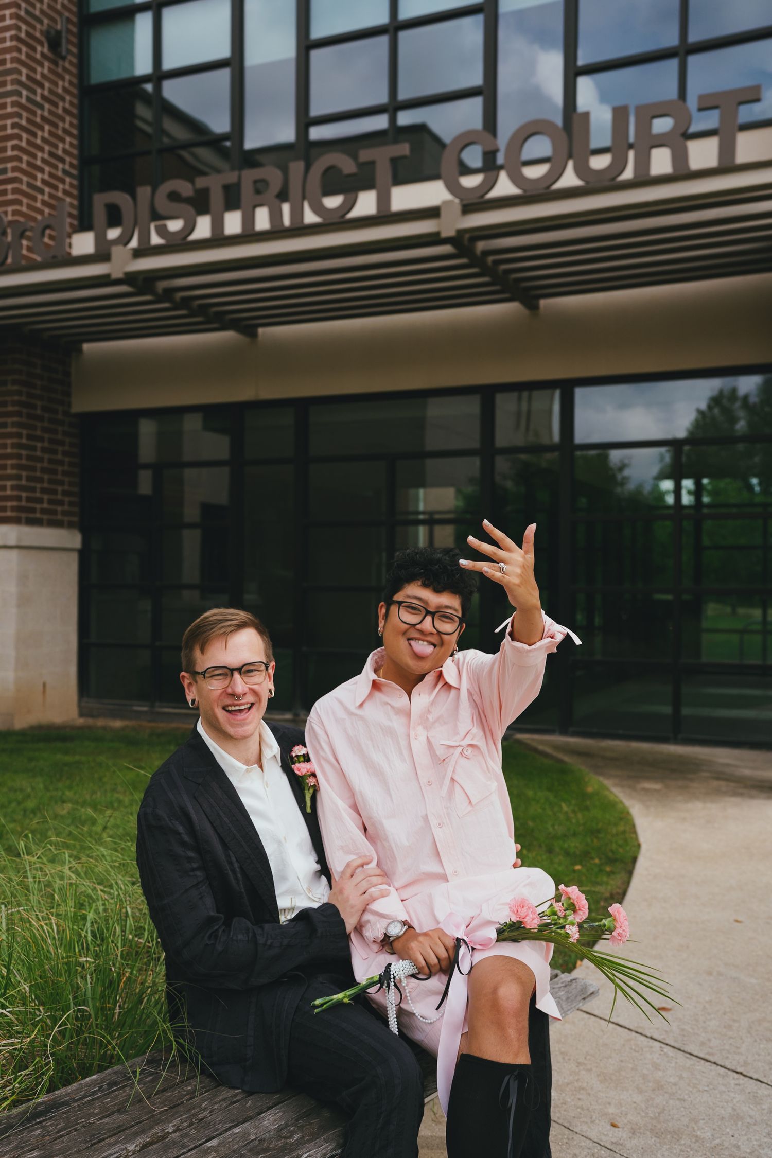 Joyful couple celebrating outside District Court, one flashing a wedding ring with pink flowers.
