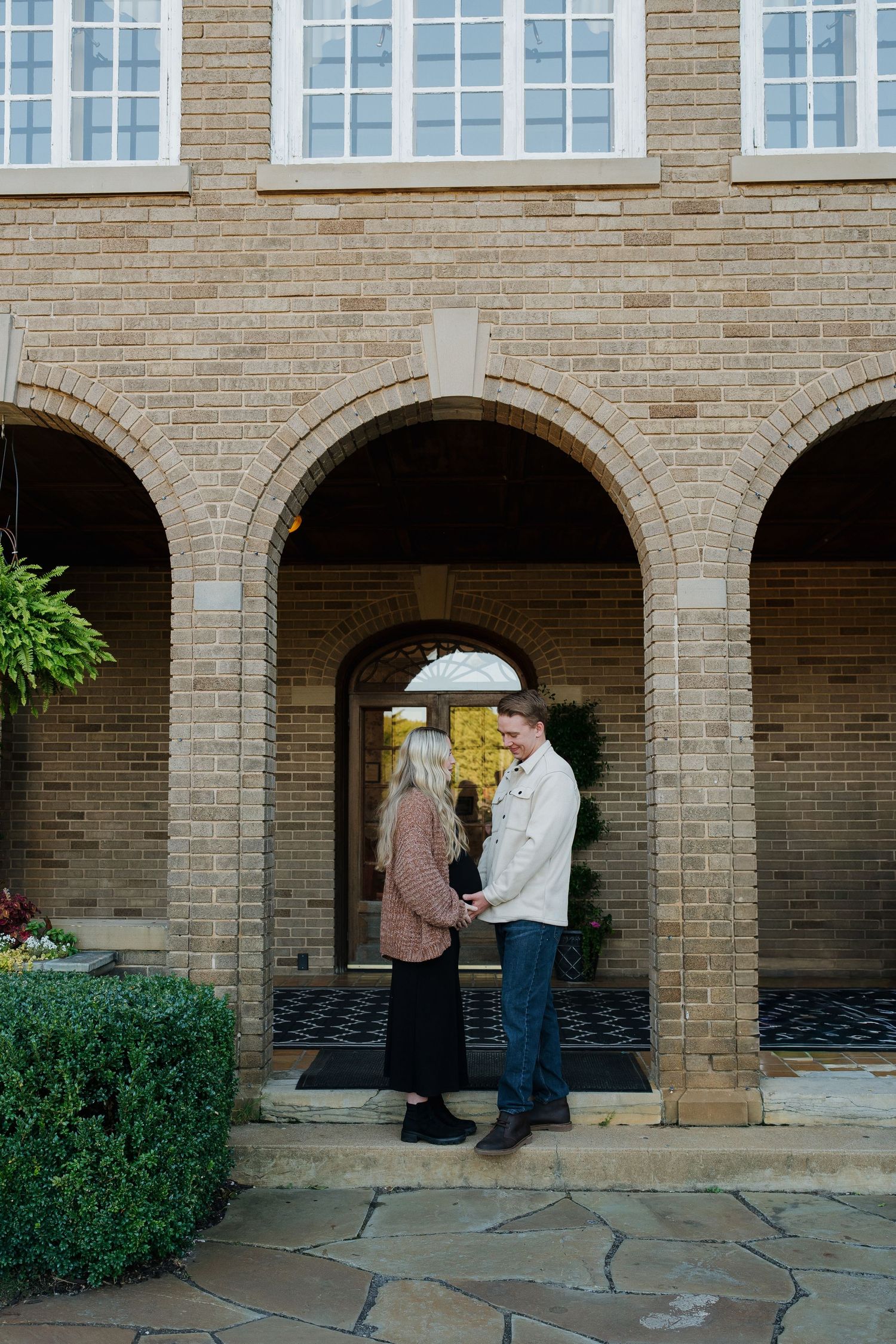 A couple holds hands facing each other outside an elegant brick building with arched windows.