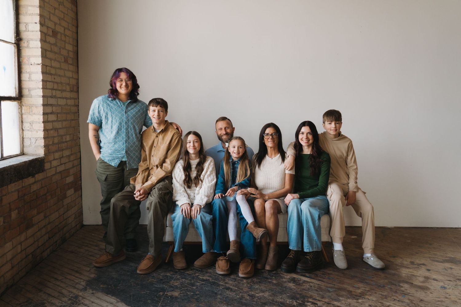 A large family of eight smiles together on a bench in a bright studio with brick walls.