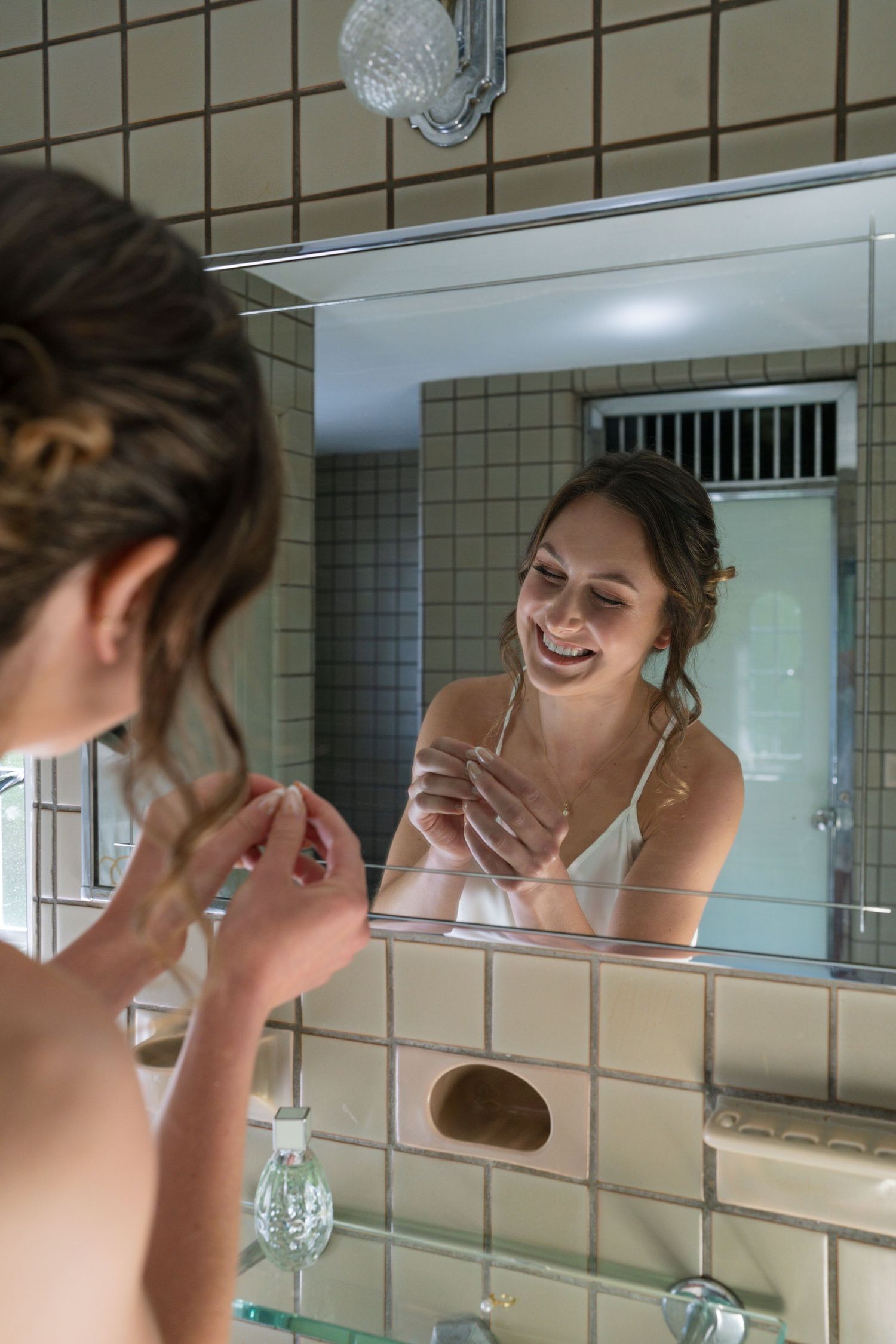 Smiling bride in white dress admires her jewelry in a bathroom mirror while getting ready.