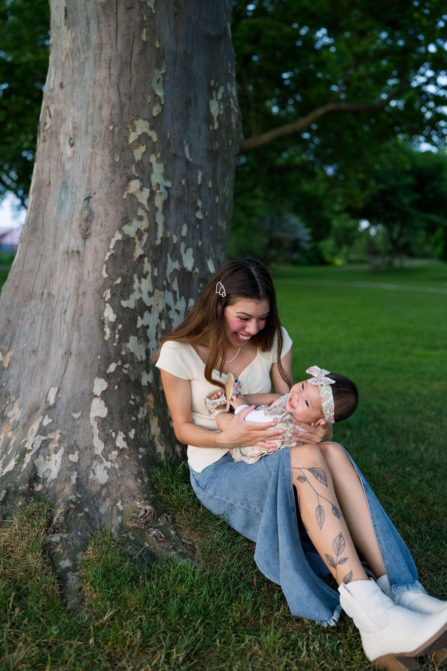 Young mother sitting against a large tree trunk cradling her newborn baby in a lush green park.