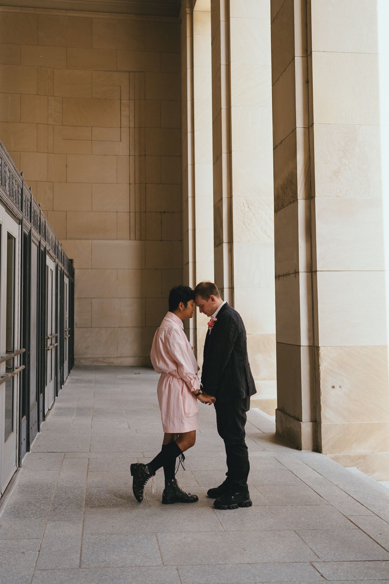 Couple holding hands facing each other in sunlit corridor, woman in pink dress and man in dark suit.
