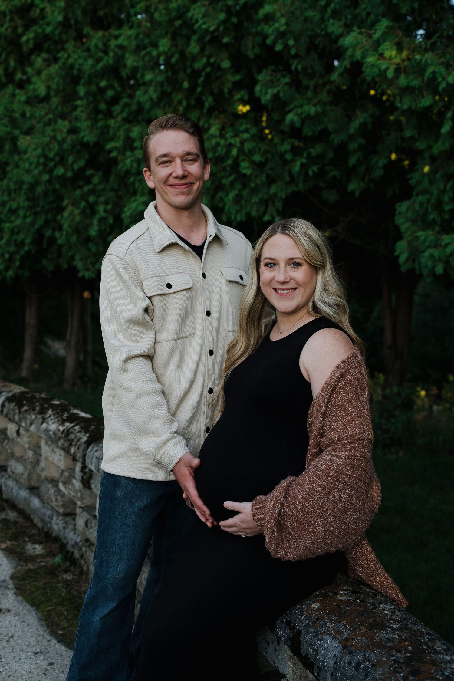 Smiling pregnant couple posing outdoors on stone bridge, both hands cradling baby bump.
