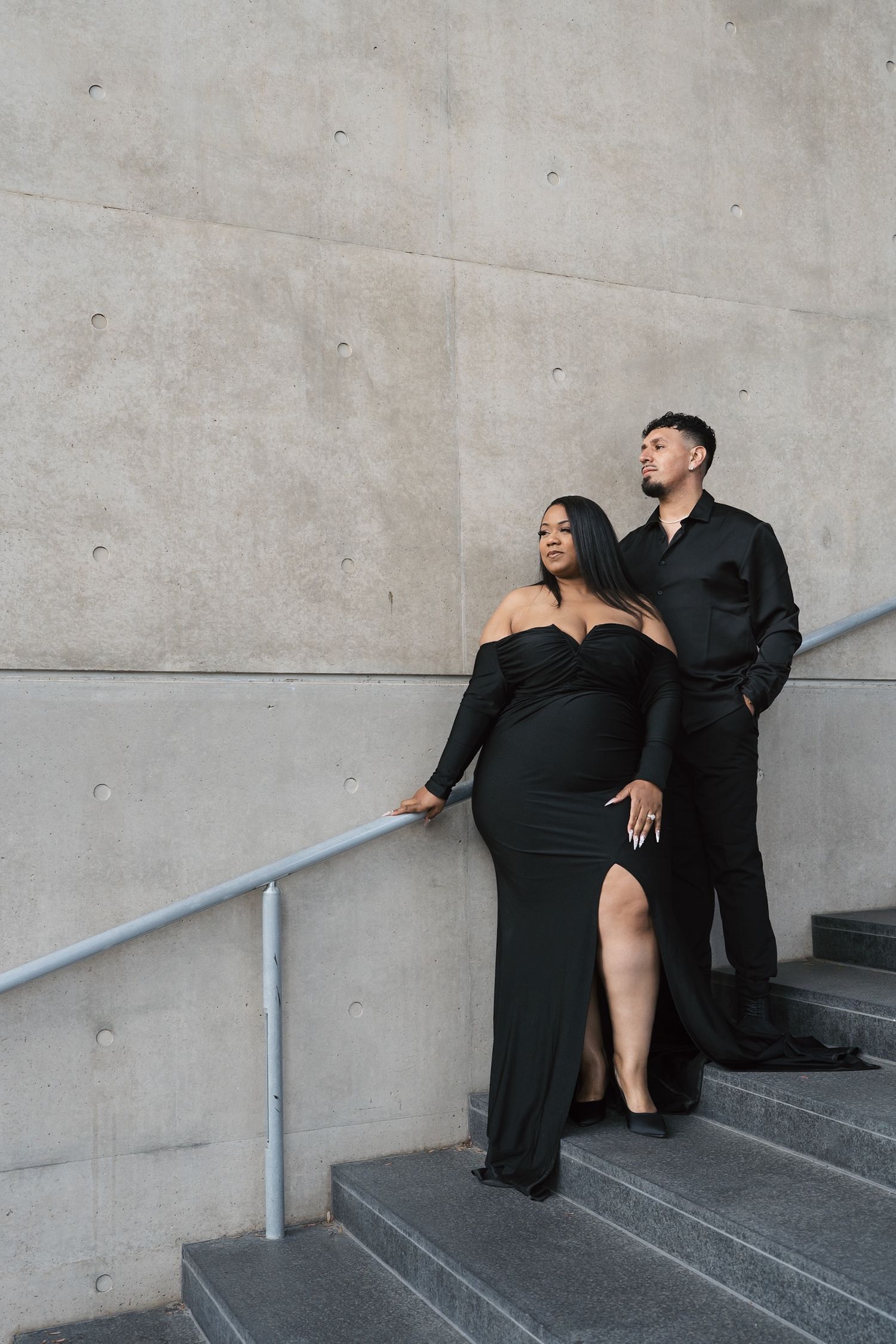 Sophisticated couple in black formal wear posing on steps against a large concrete wall.