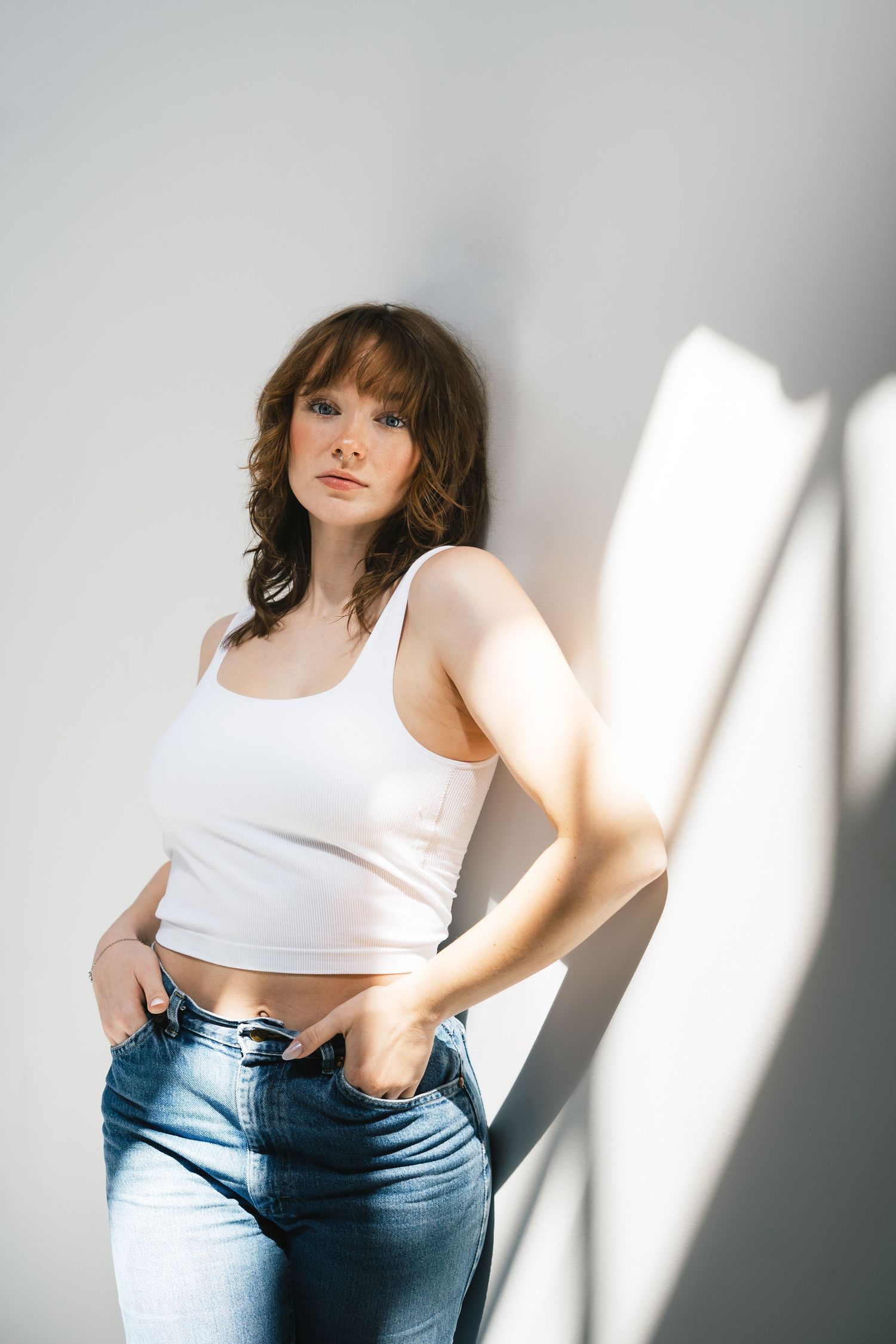Woman in white crop tank top and blue jeans posing with hands in pockets against sunlit white wall.