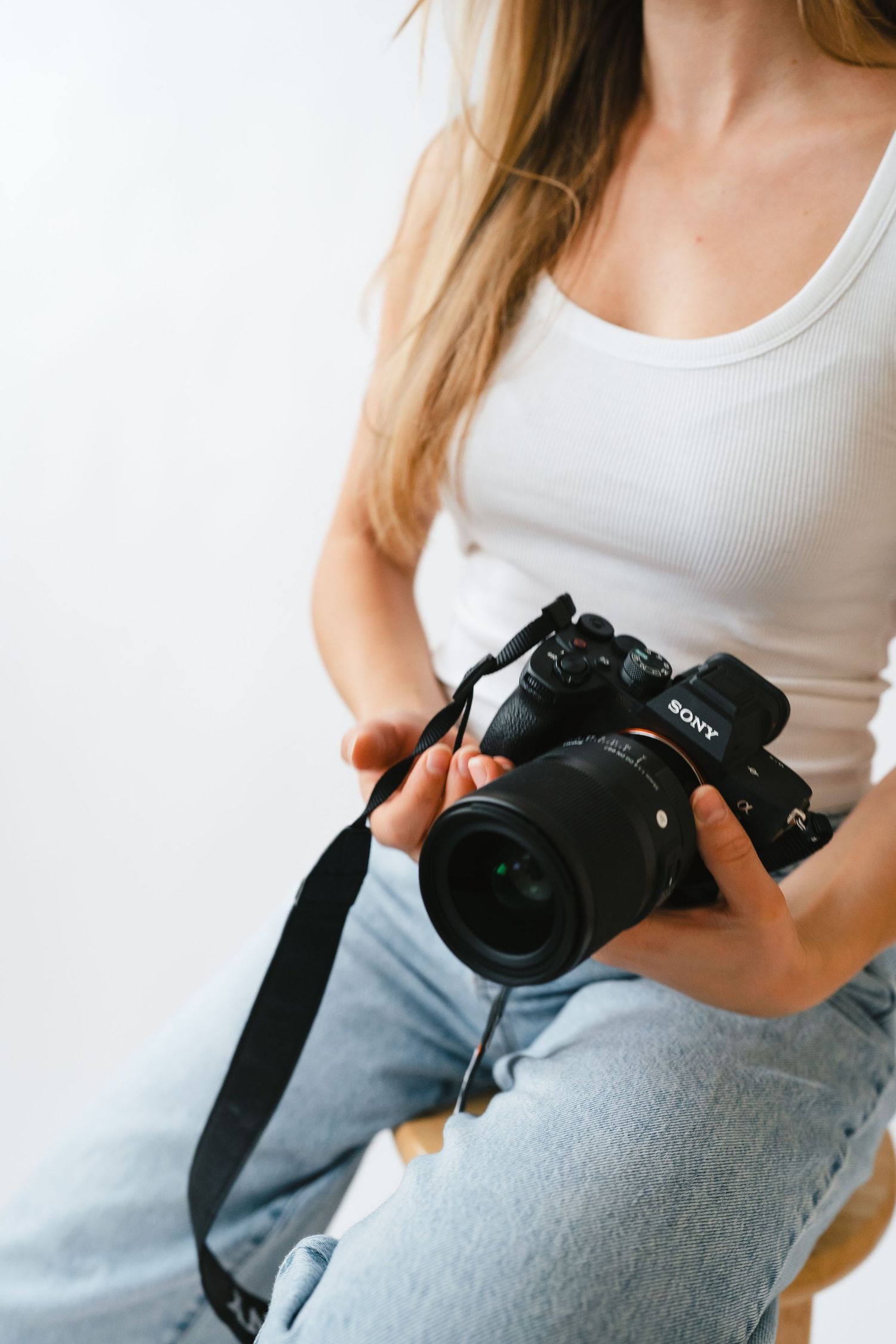 Close-up of woman in white tank top and jeans holding a Sony mirrorless camera with black strap.