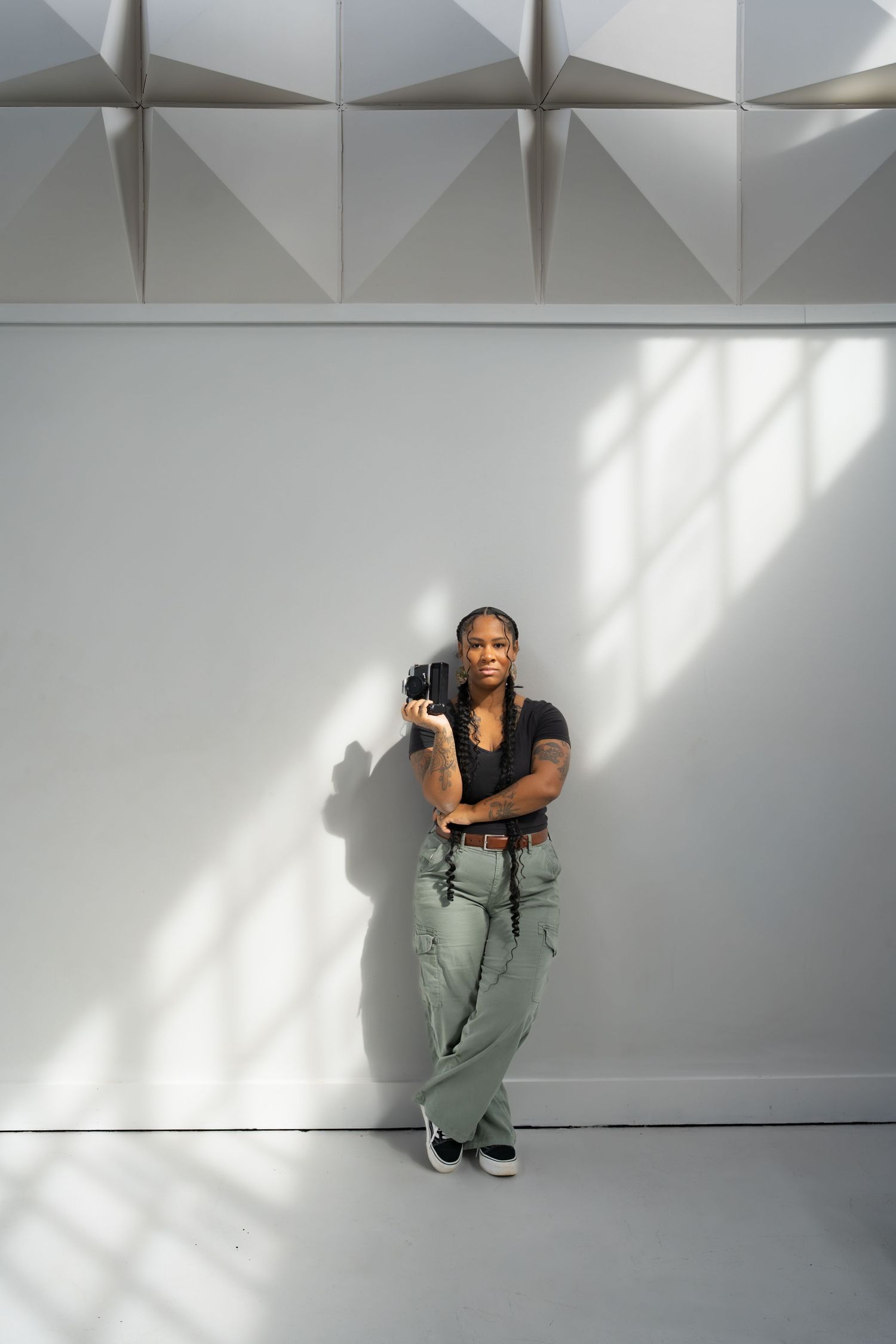 A Black woman with braids leans against a white wall holding a film camera, wearing a black top and sage green cargo pants.