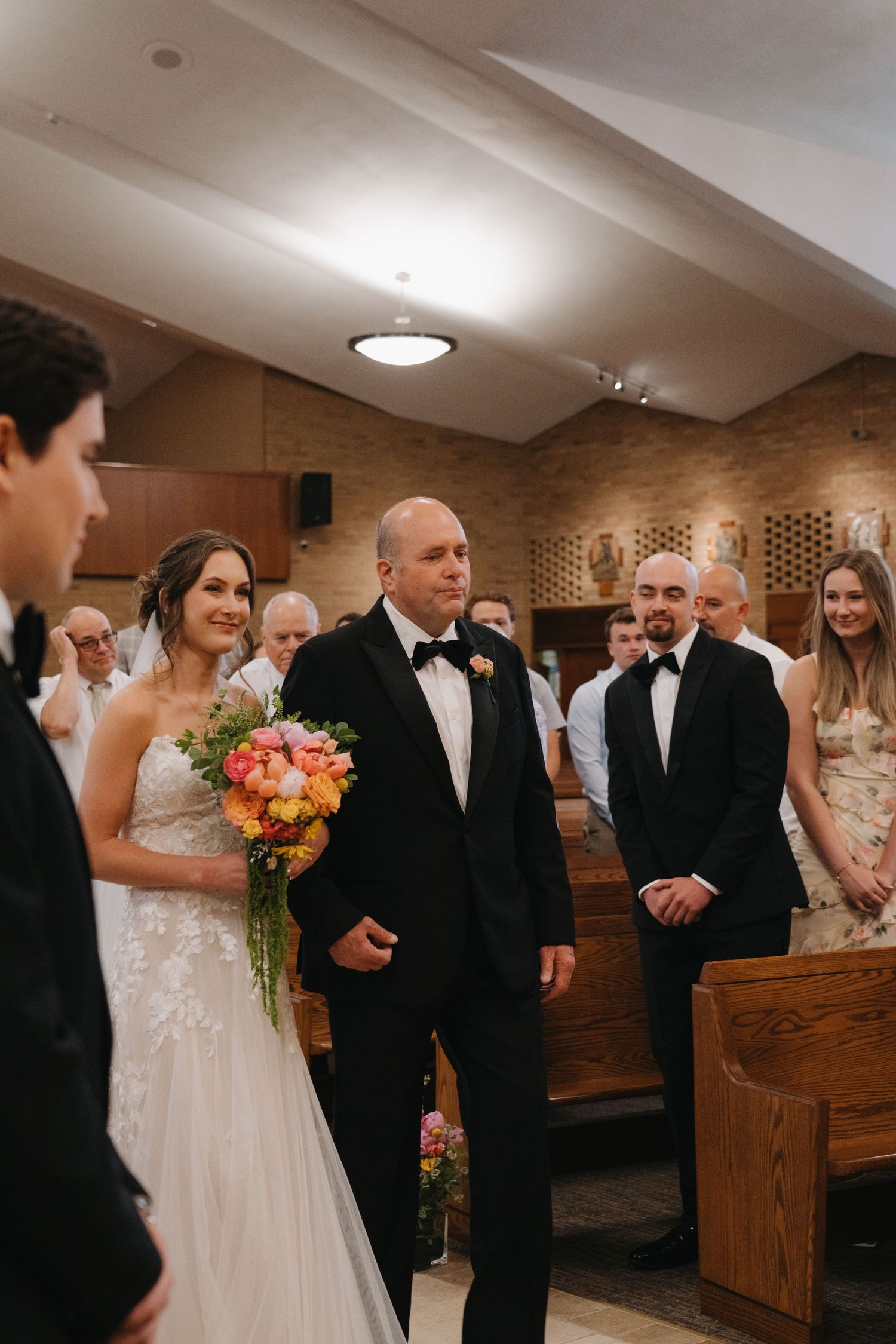 Father escorts smiling bride down the church aisle as groom and guests watch the wedding processional.