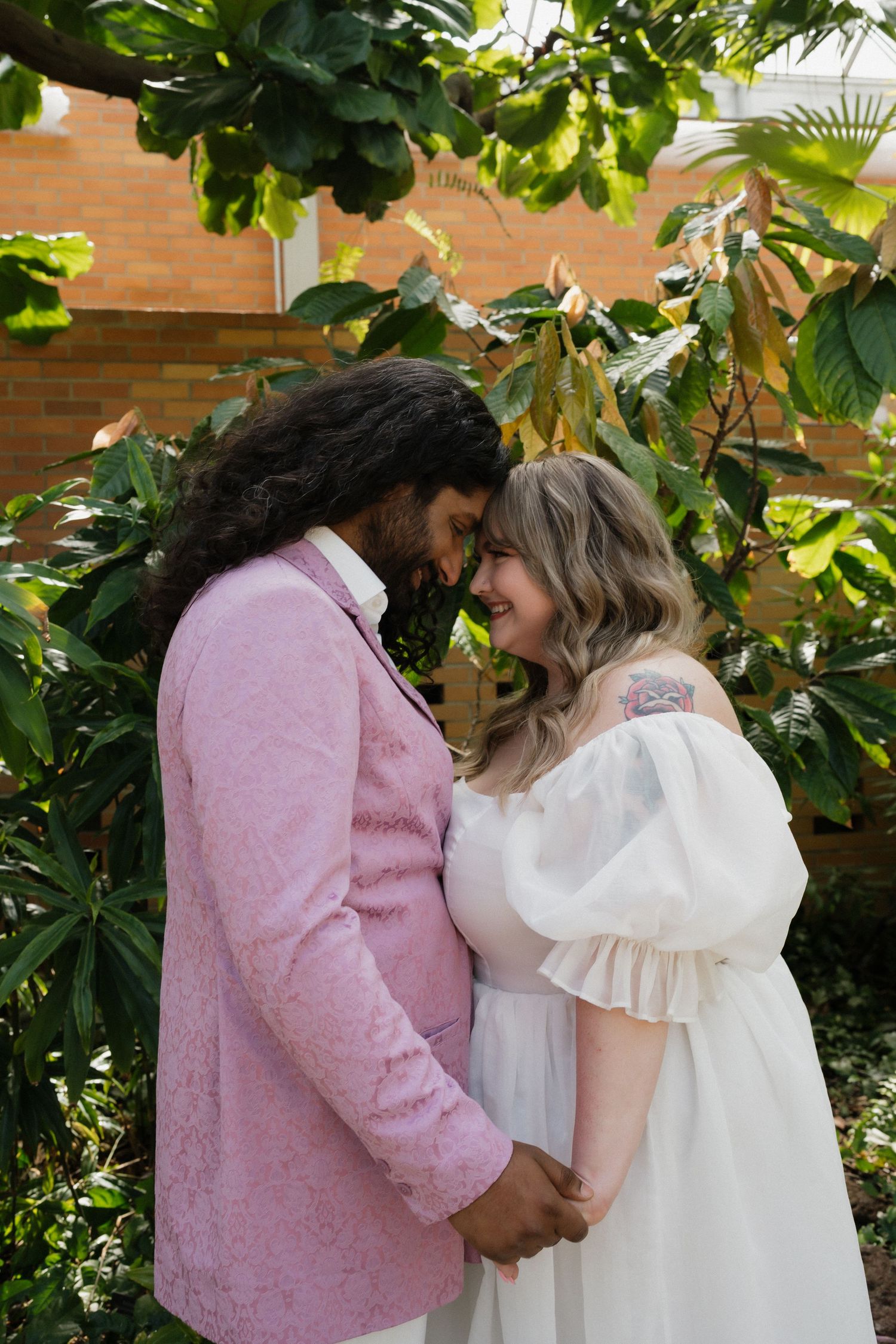 Interracial wedding couple holding hands and smiling face to face among green plants.
