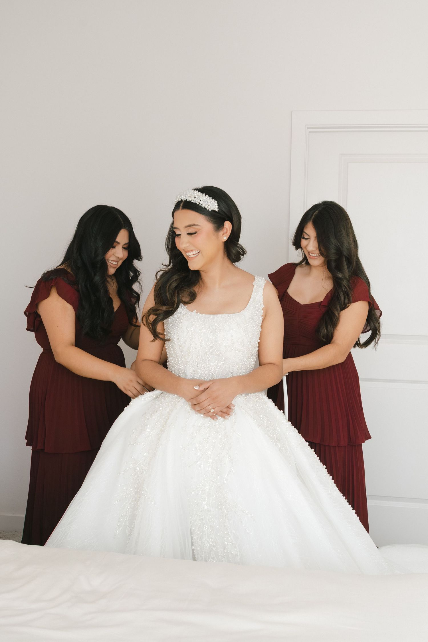Bride in pearl-embellished white ball gown smiling as two bridesmaids in burgundy dresses assist her.