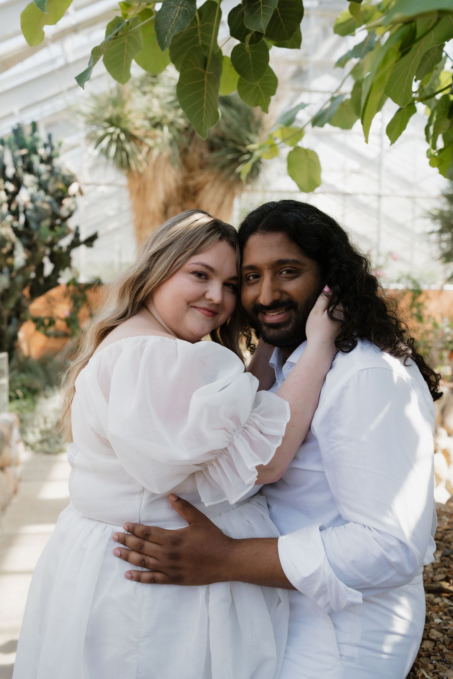 Interracial couple embracing and smiling in a bright greenhouse surrounded by tropical plants.