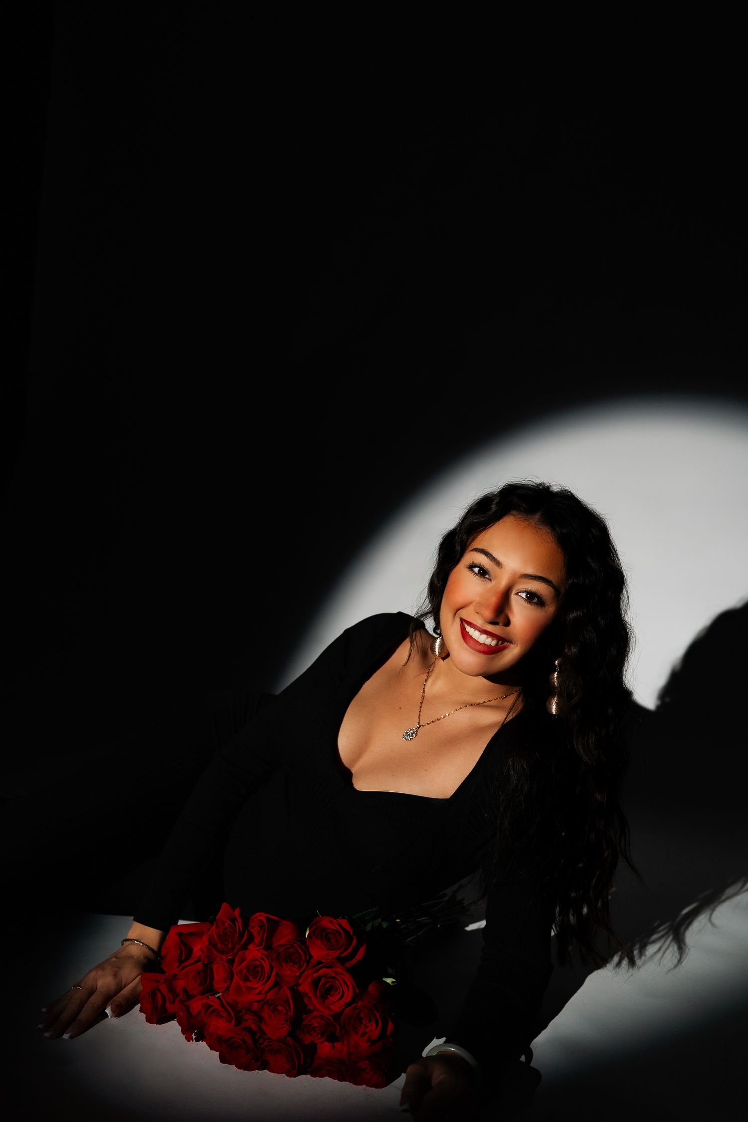 Glamorous woman in black outfit smiling with red roses bouquet in a dramatic spotlight portrait.