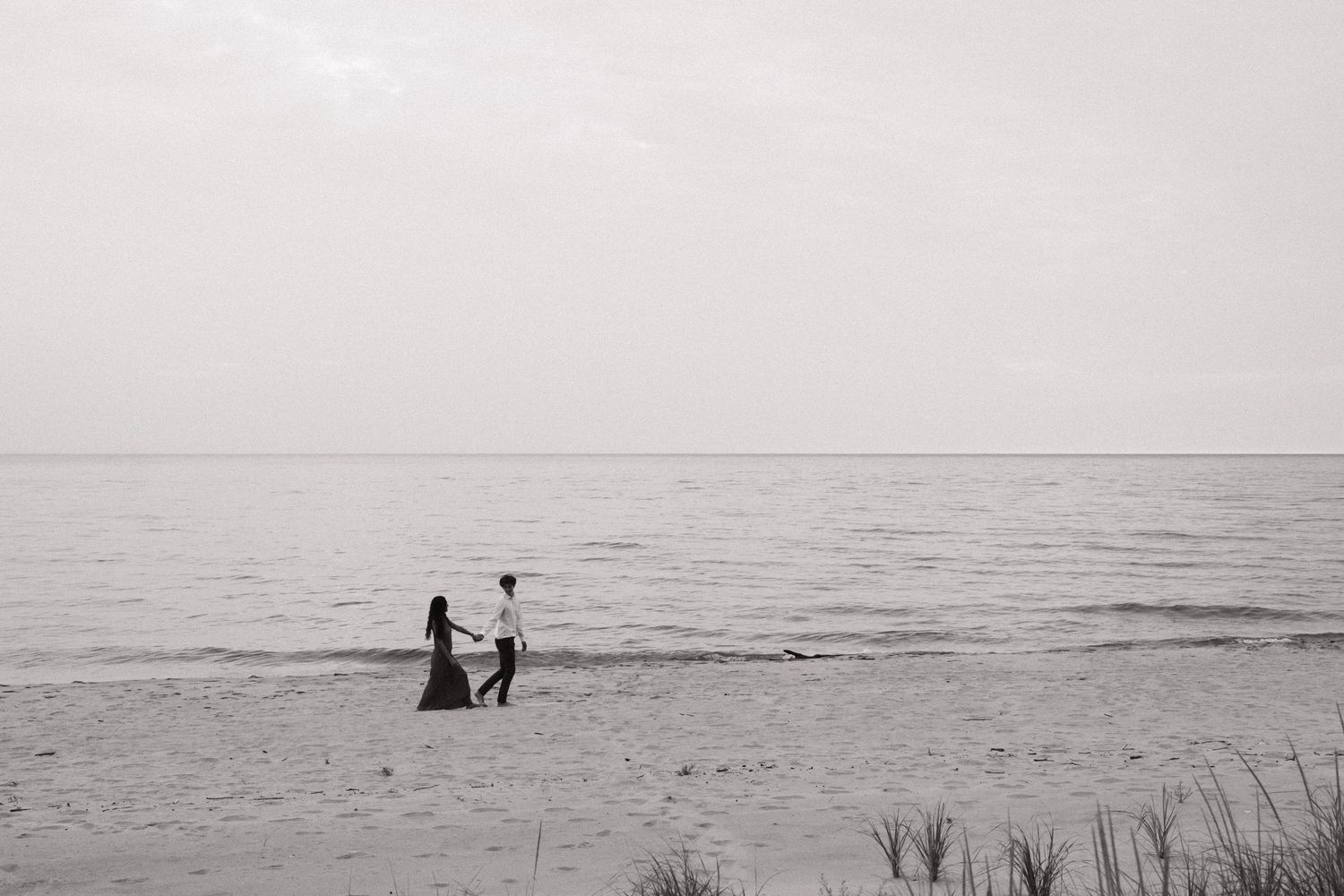 Black and white photo of a couple holding hands on a serene beach with calm ocean waters in the background.