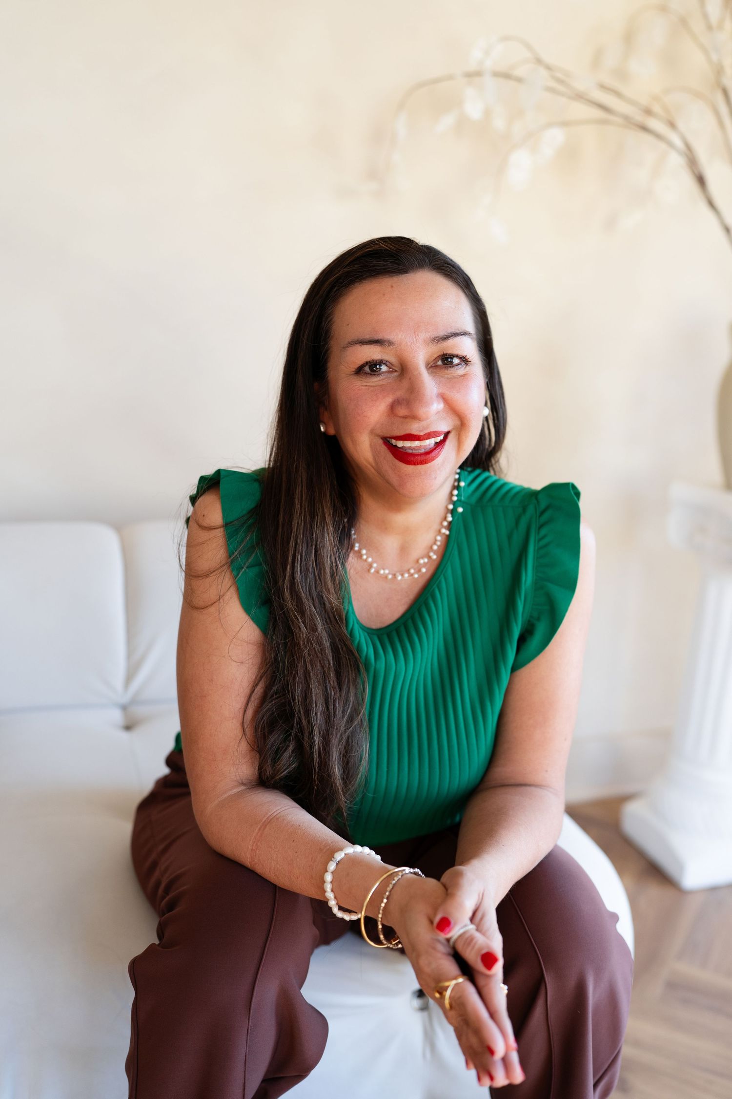Smiling Latina woman in a green top and brown pants sitting on a white couch indoors.