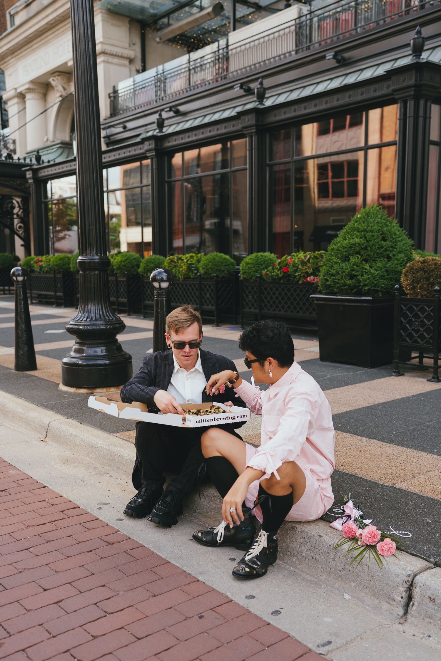 Newlywed couple in sunglasses eating pizza on urban sidewalk after courthouse elopement.