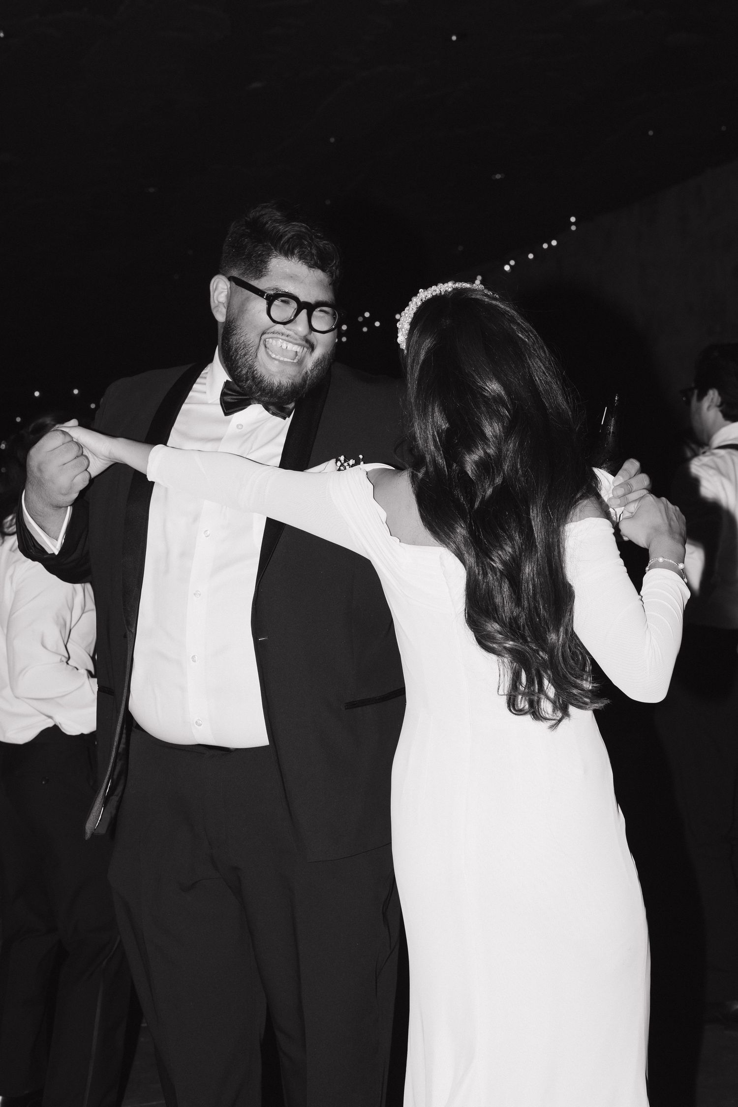 Black and white photo of a smiling groom in tuxedo dancing with bride in white gown at wedding reception.