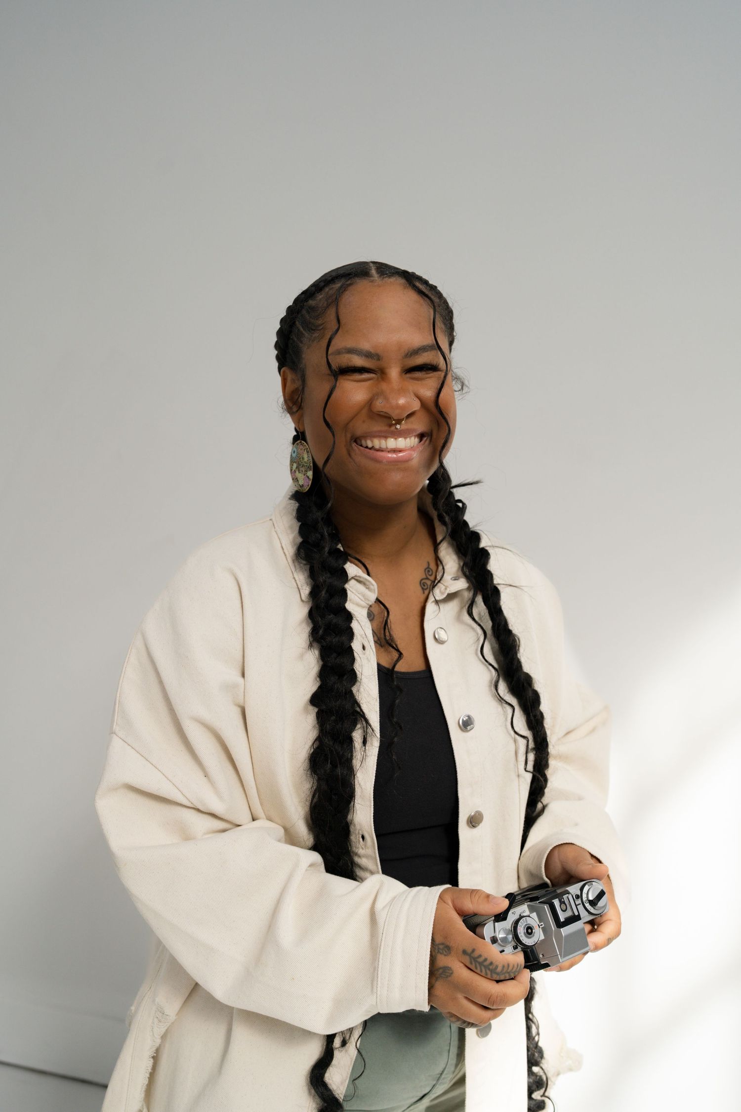 A smiling Black woman with long braids holds a film camera, wearing a white jacket over a black top in a bright studio.