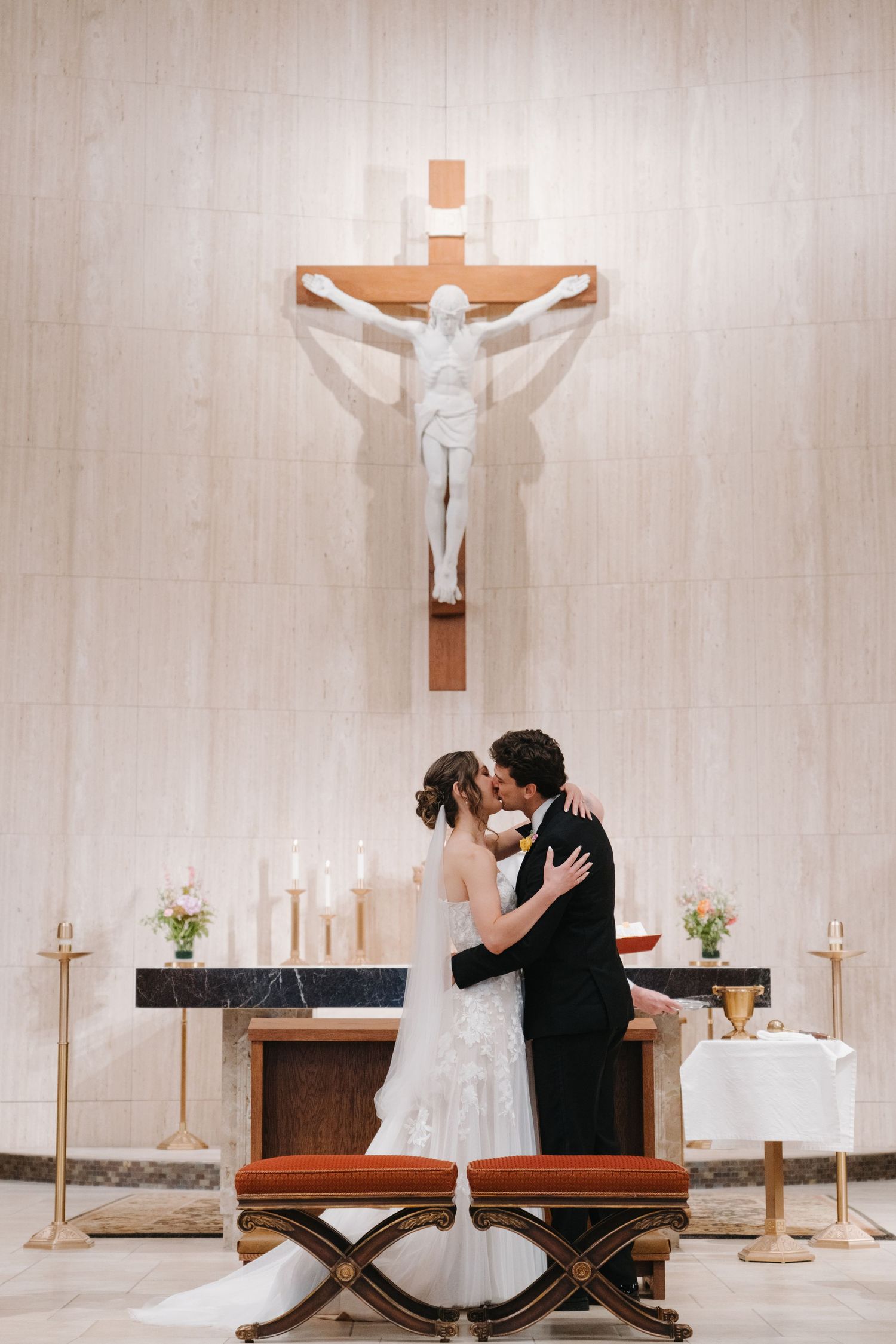Newlyweds share first kiss at church altar beneath a crucifix surrounded by candles and flowers.