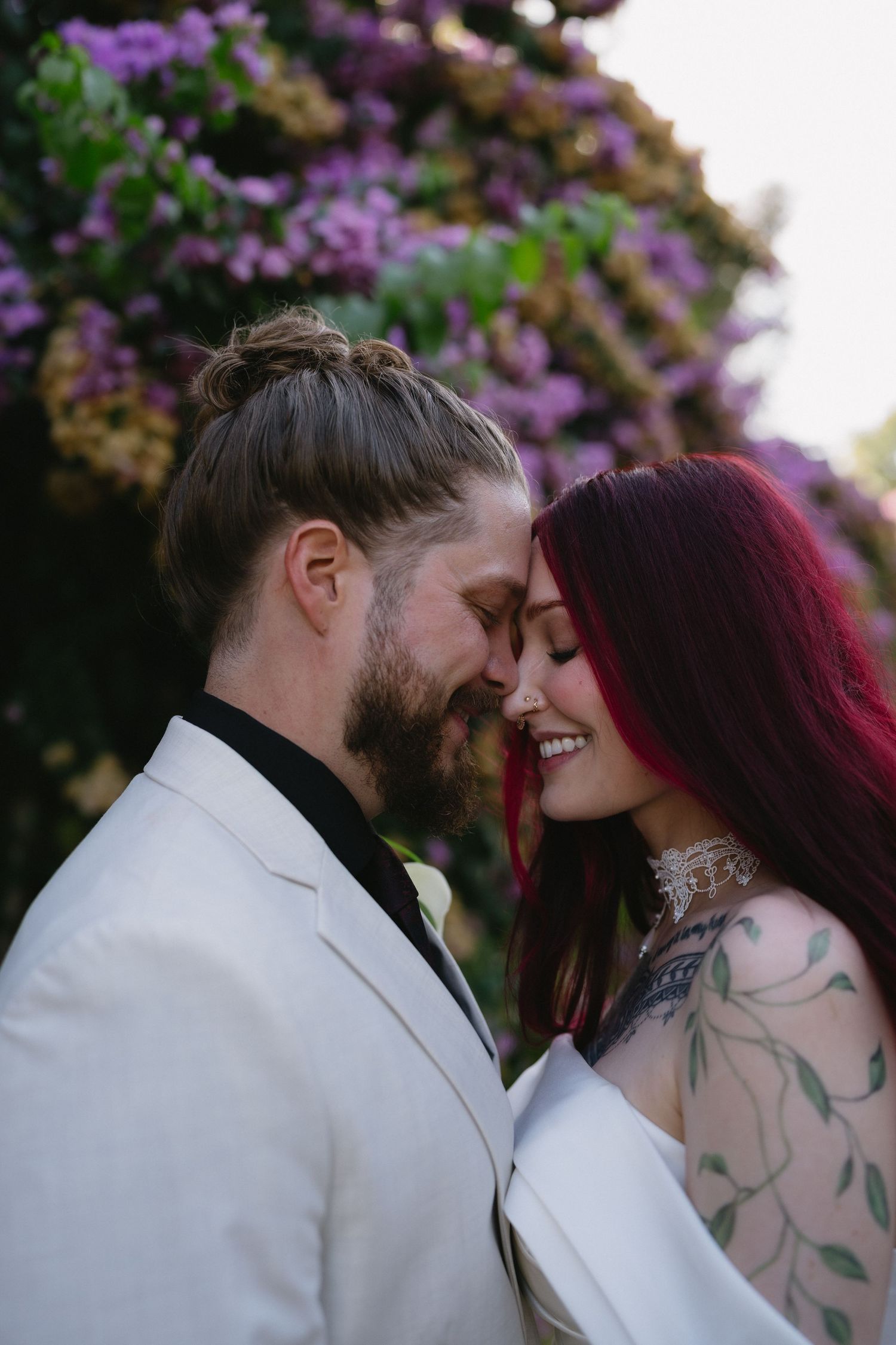 A bearded groom in a white suit and red-haired bride with tattoos share a romantic moment among purple blooming flowers.