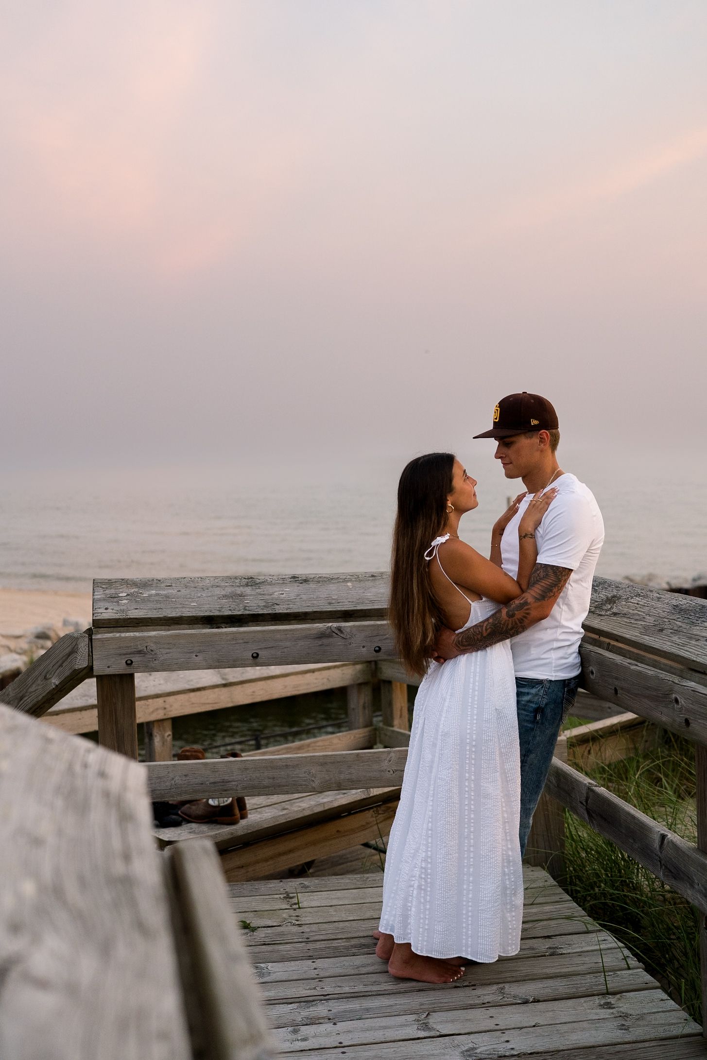 Young couple holding each other on wooden beach boardwalk at dusk, woman in white dress, man in white tee.