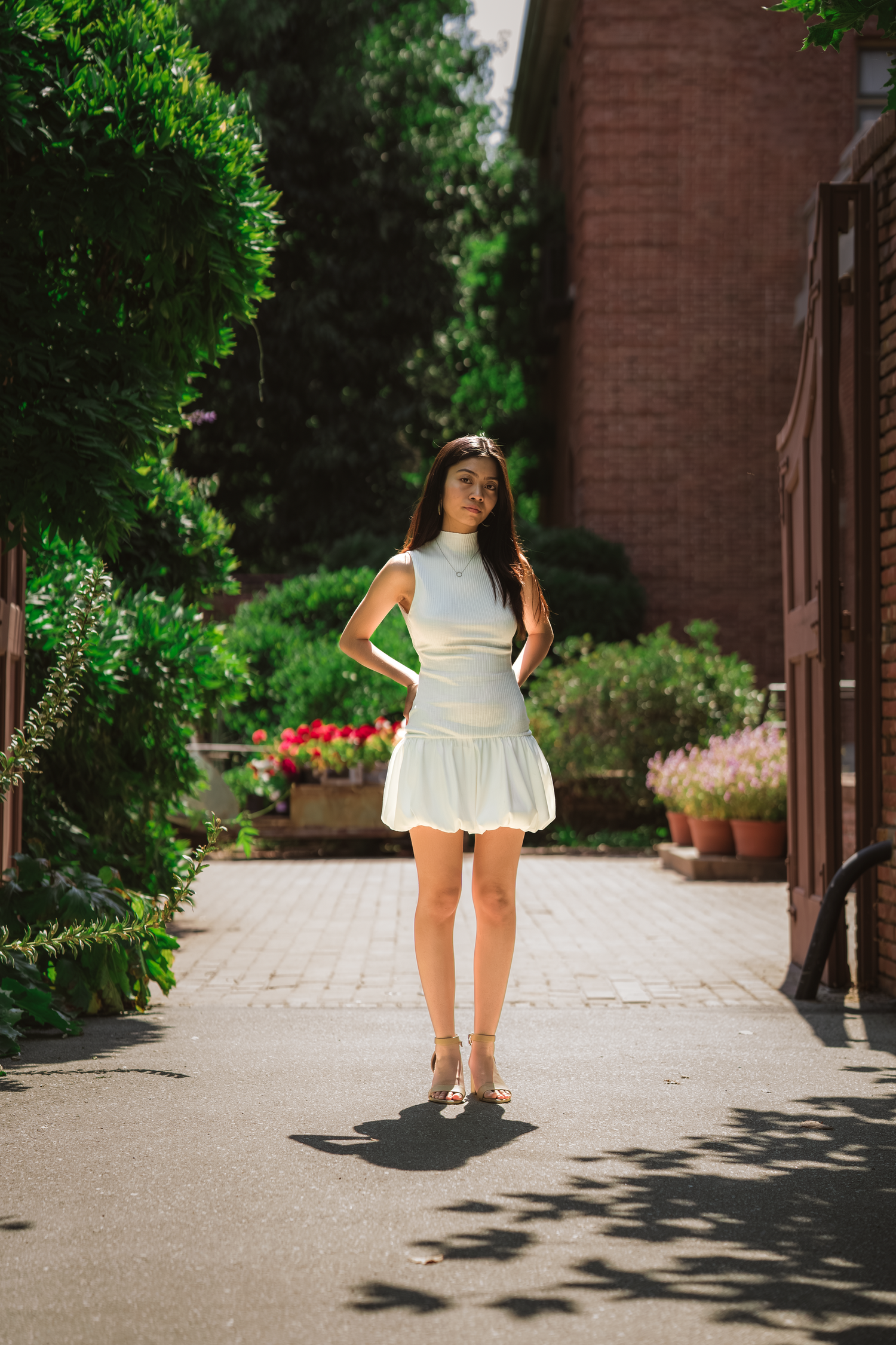 Young woman in white mini dress poses confidently in a lush green garden courtyard.