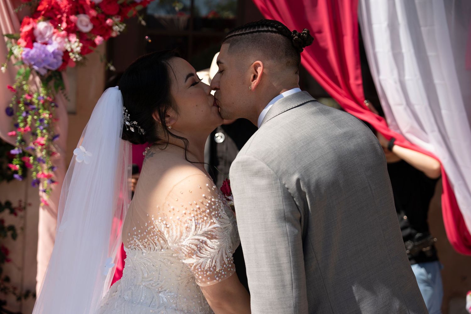 A bride and groom share their first kiss at a floral wedding altar with red draping and colorful flowers.