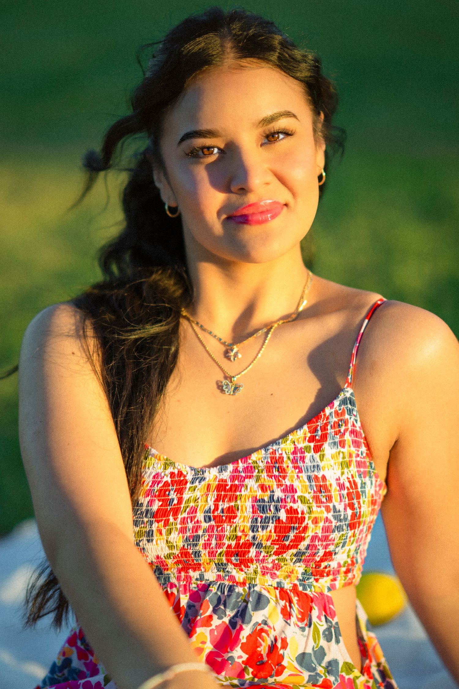 Young woman in floral sundress smiling outdoors in golden sunlight, wearing layered necklaces.