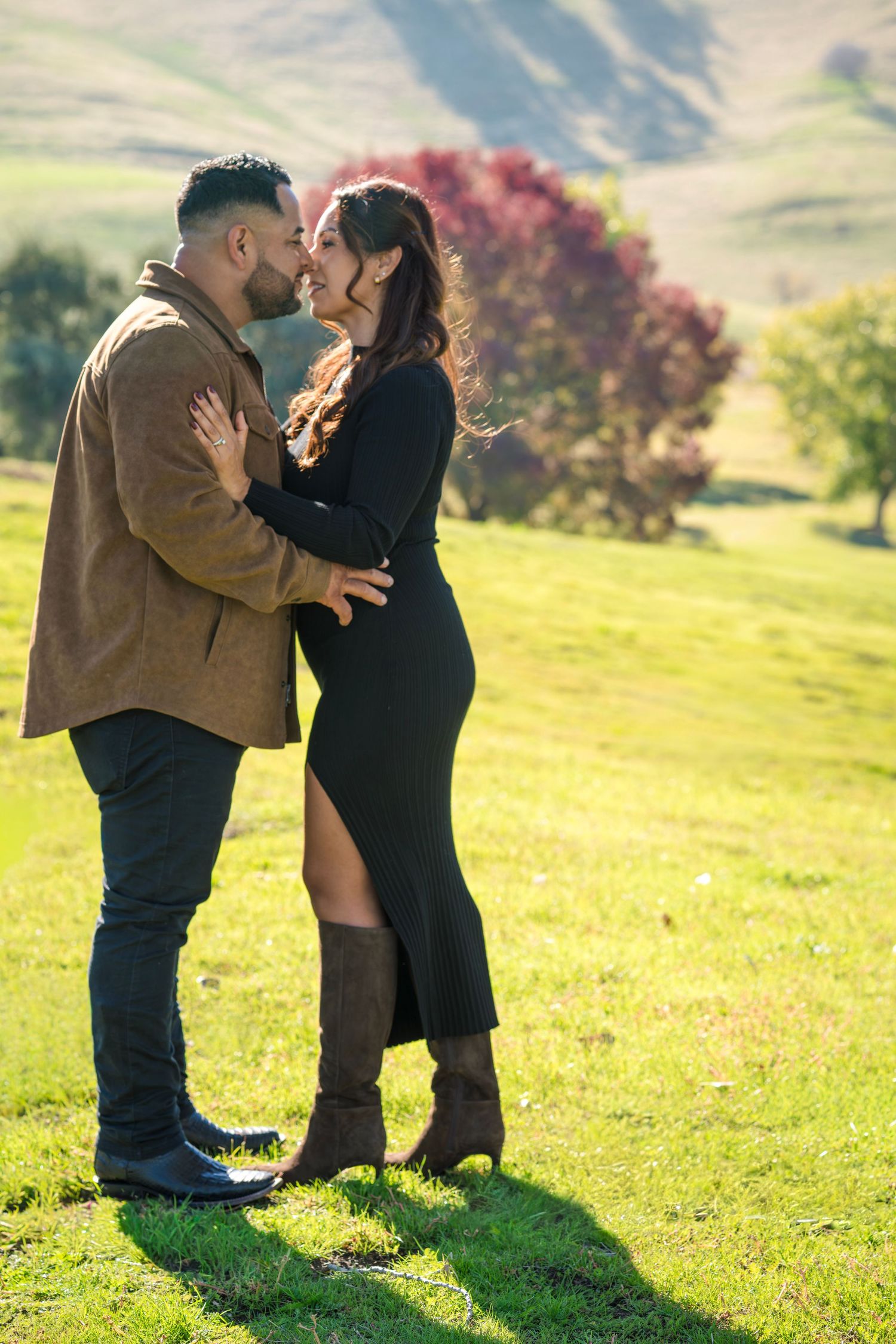 Couple sharing a romantic almost-kiss embrace in a sunny green meadow with autumn trees in the background.