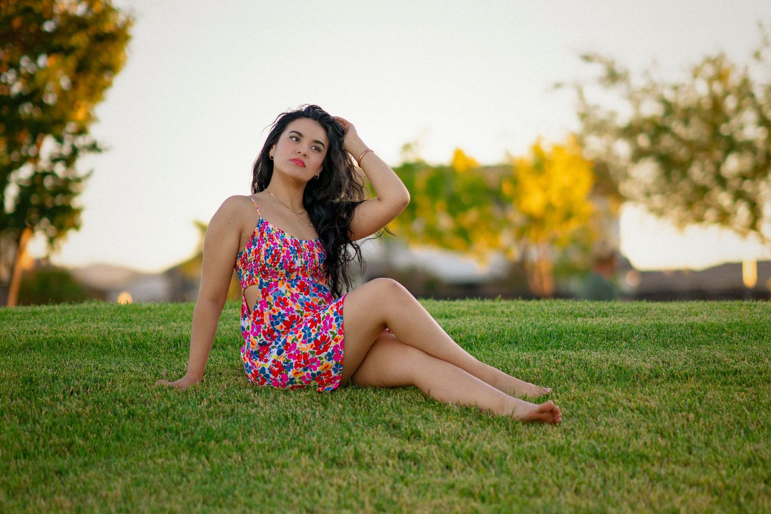 Young woman in floral sundress sitting gracefully on lush green grass outdoors during golden hour.