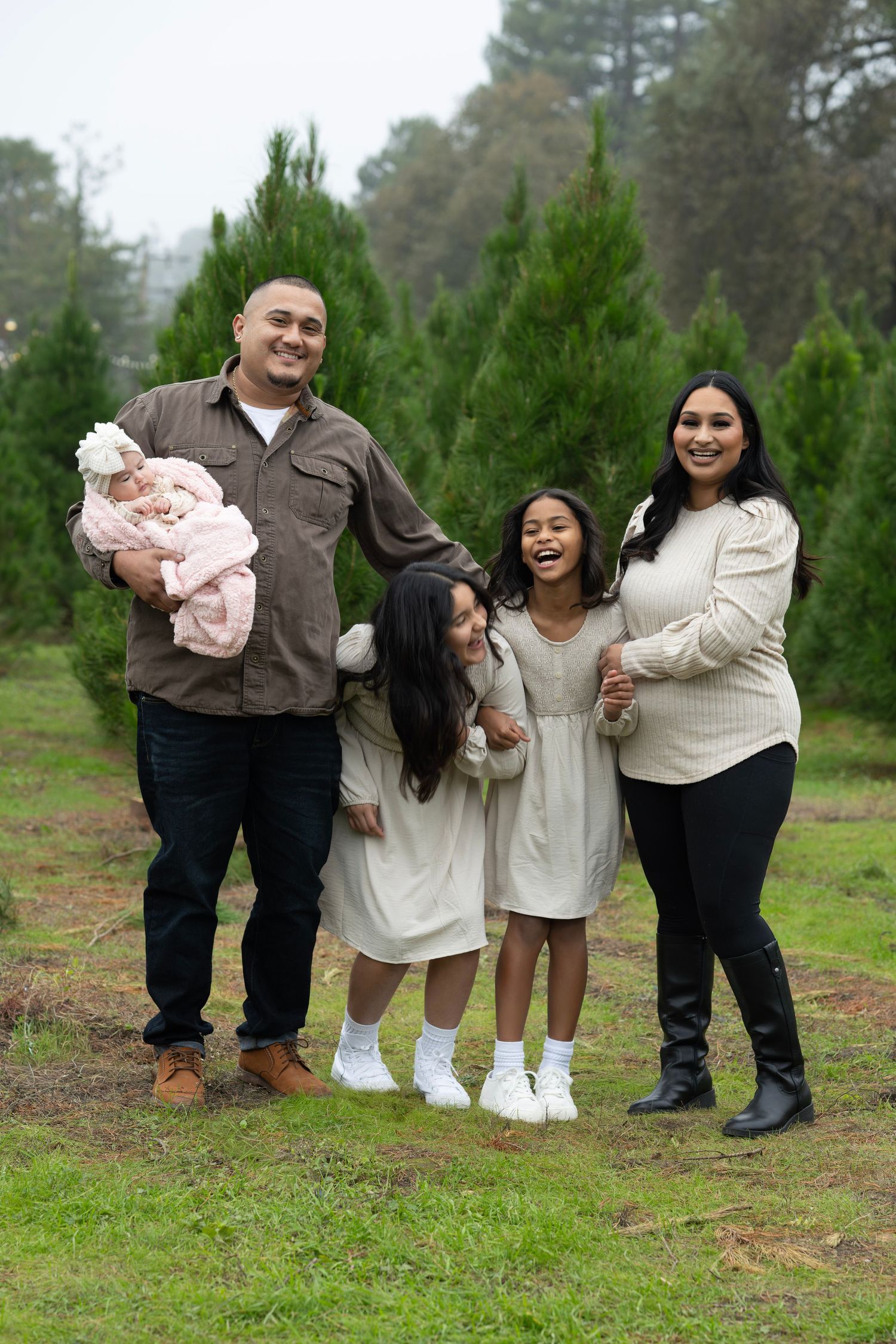 Happy family of five posing among tall green Christmas trees on a misty day.