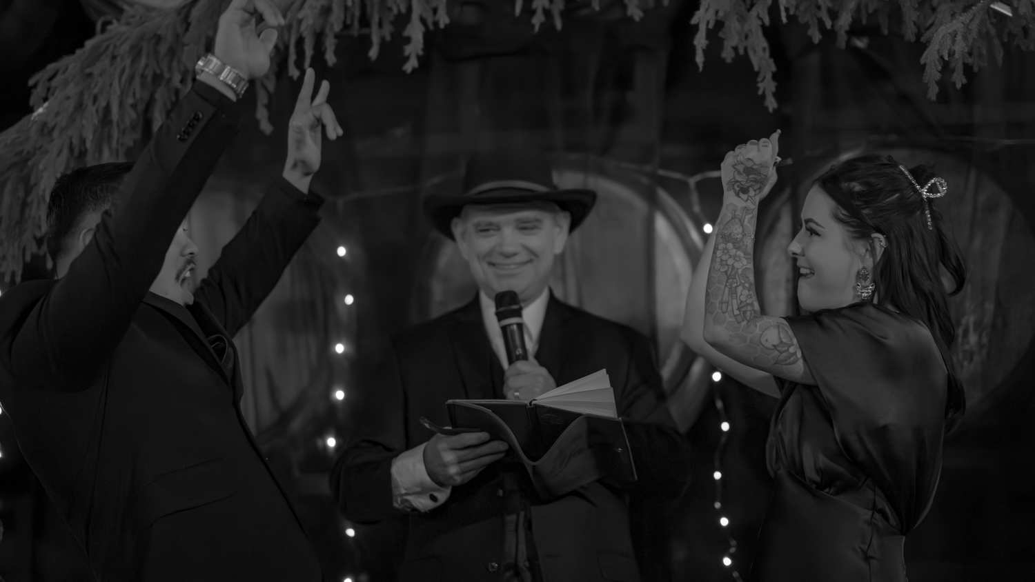 Black and white photo of couple raising hands during wedding ceremony with officiant holding papers indoors.