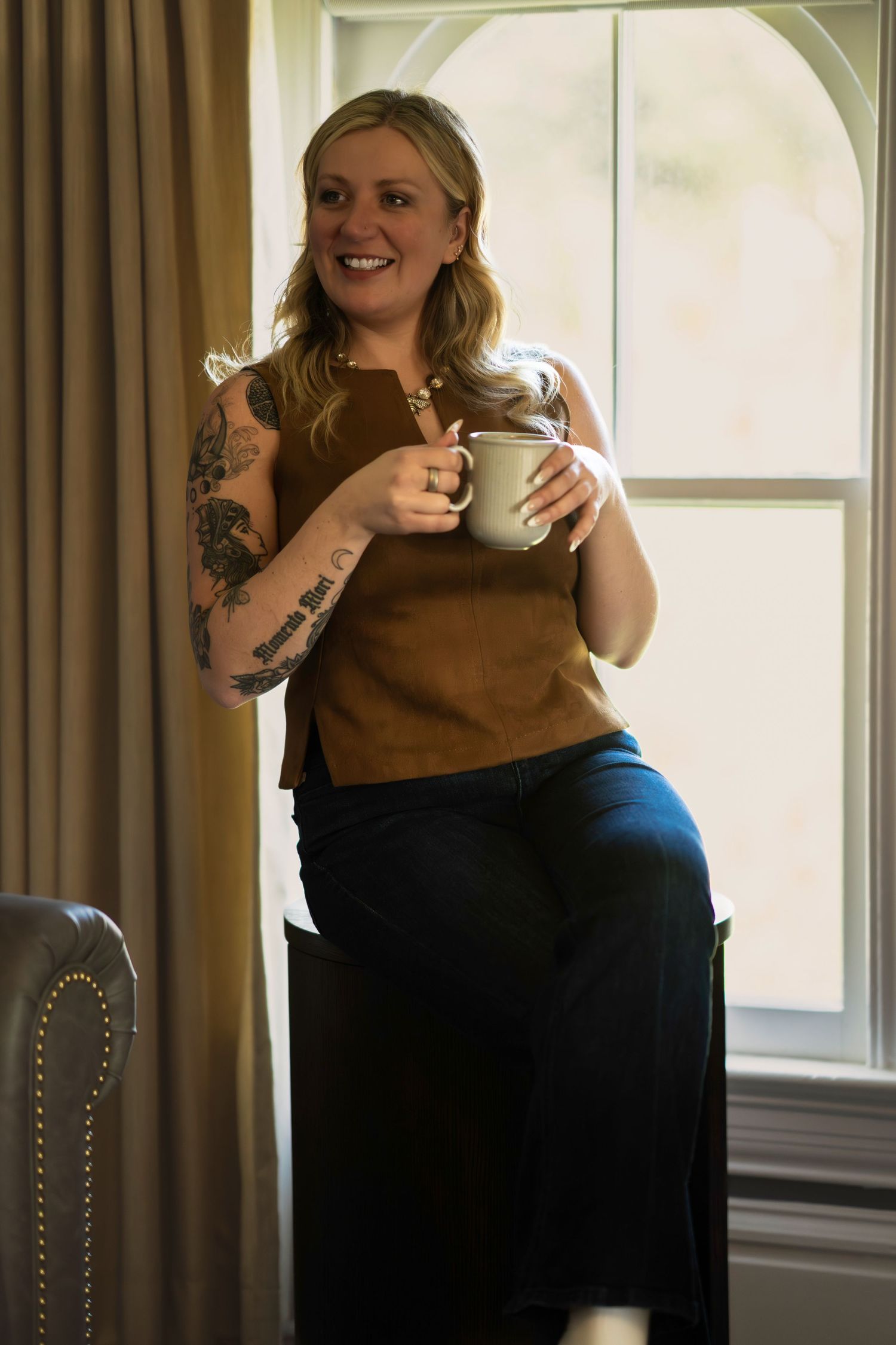 Smiling blonde woman with tattoos holding a white mug, wearing brown top and navy pants, standing by a bright window.