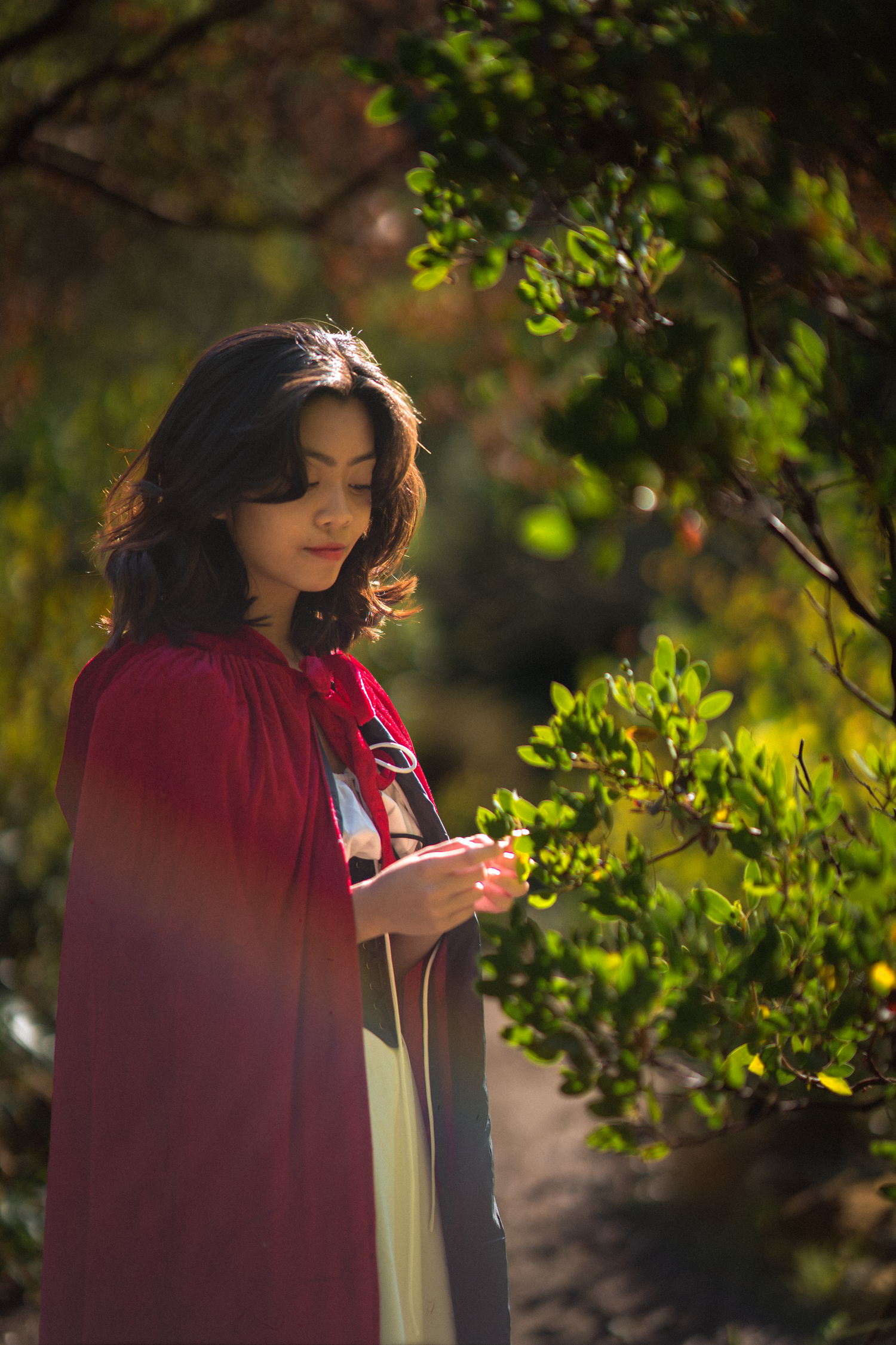 A young woman in a red cape and cream dress stands in a sunlit garden, gently touching green leaves.