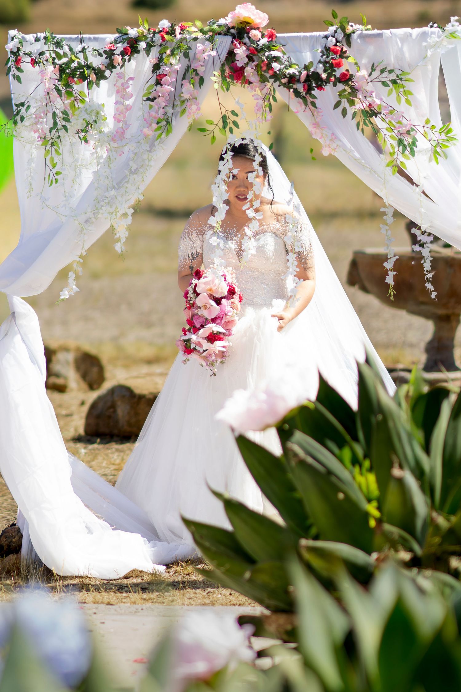 Bride in white lace gown holding pink bouquet stands under floral arch with white draping outdoors.