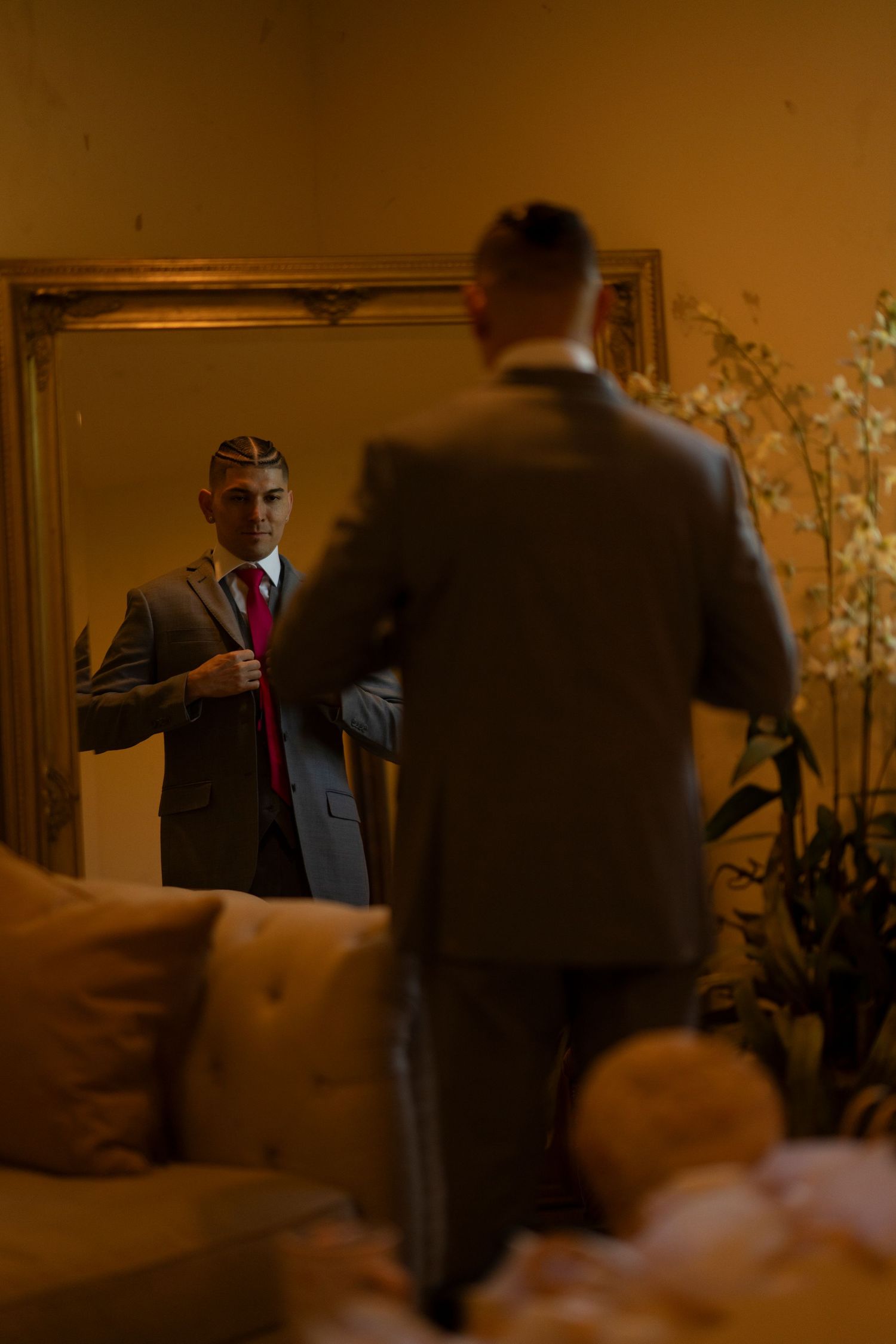 A well-dressed man in a gray suit adjusts his red tie while standing before a gold-framed mirror in a warmly lit room.