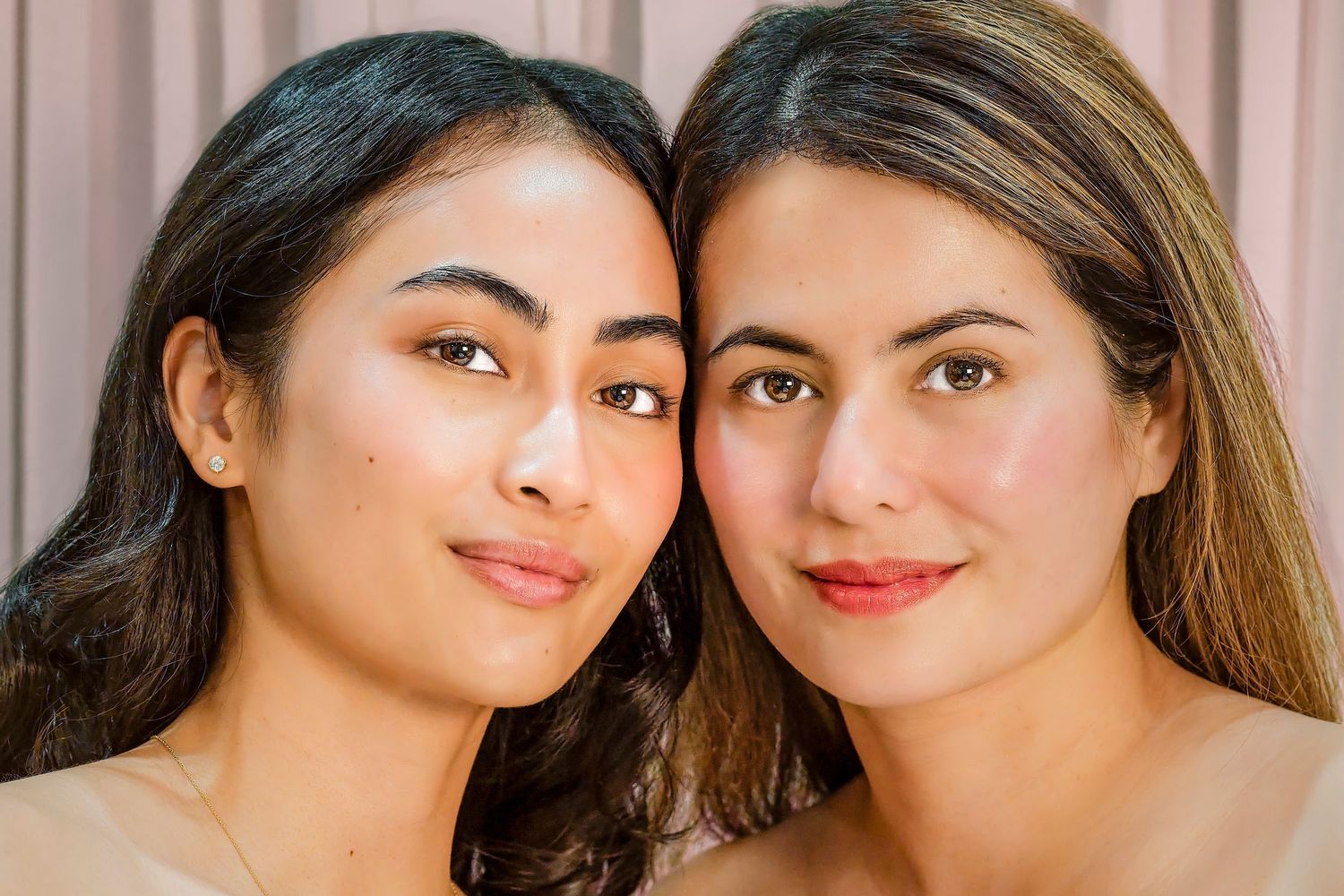 Two young women with glowing skin pose cheek to cheek, smiling softly against a neutral pink background.
