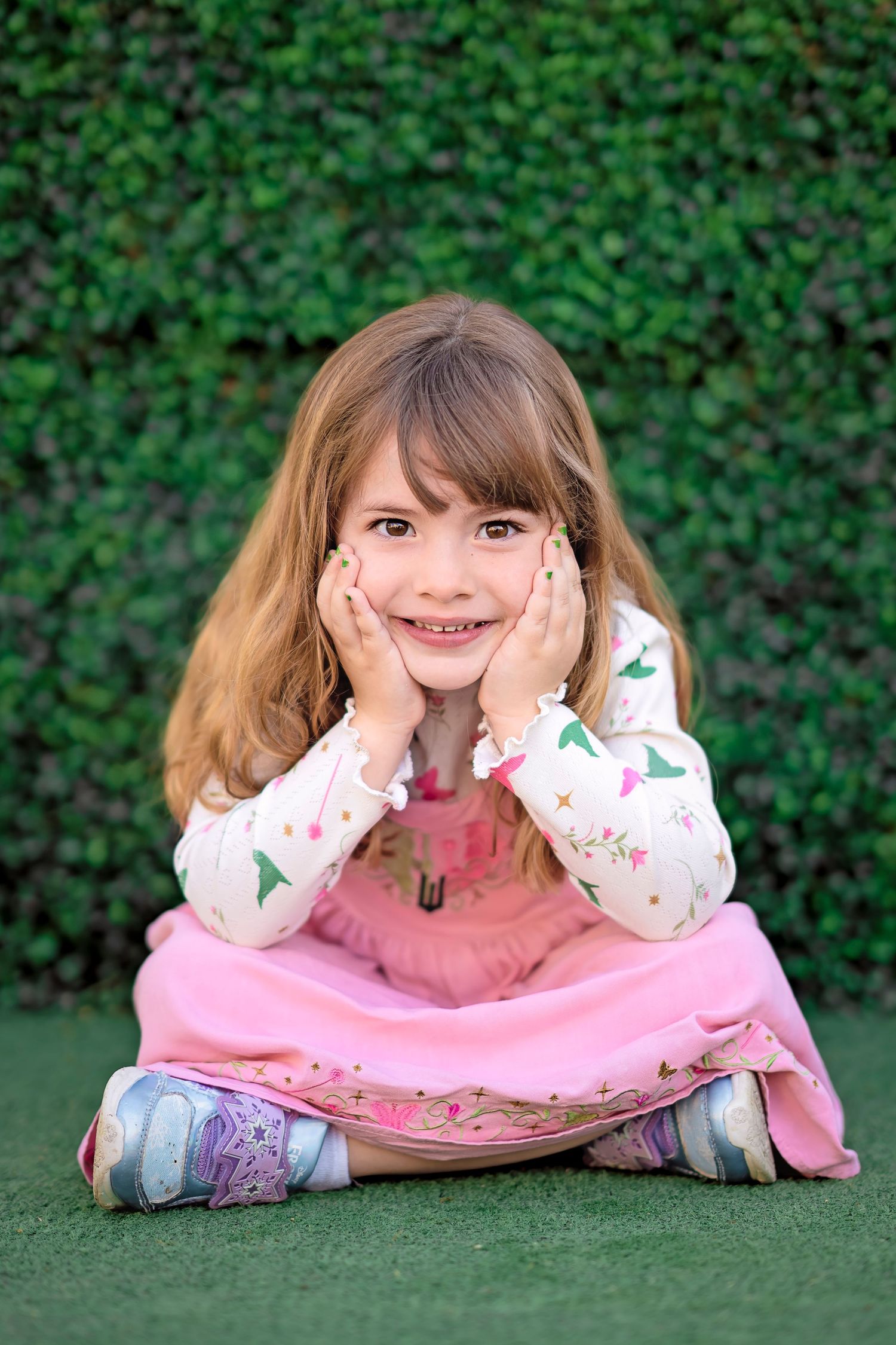 A smiling little girl in a pink dress sits cross-legged on grass with hands on cheeks, posing in front of a green hedge.