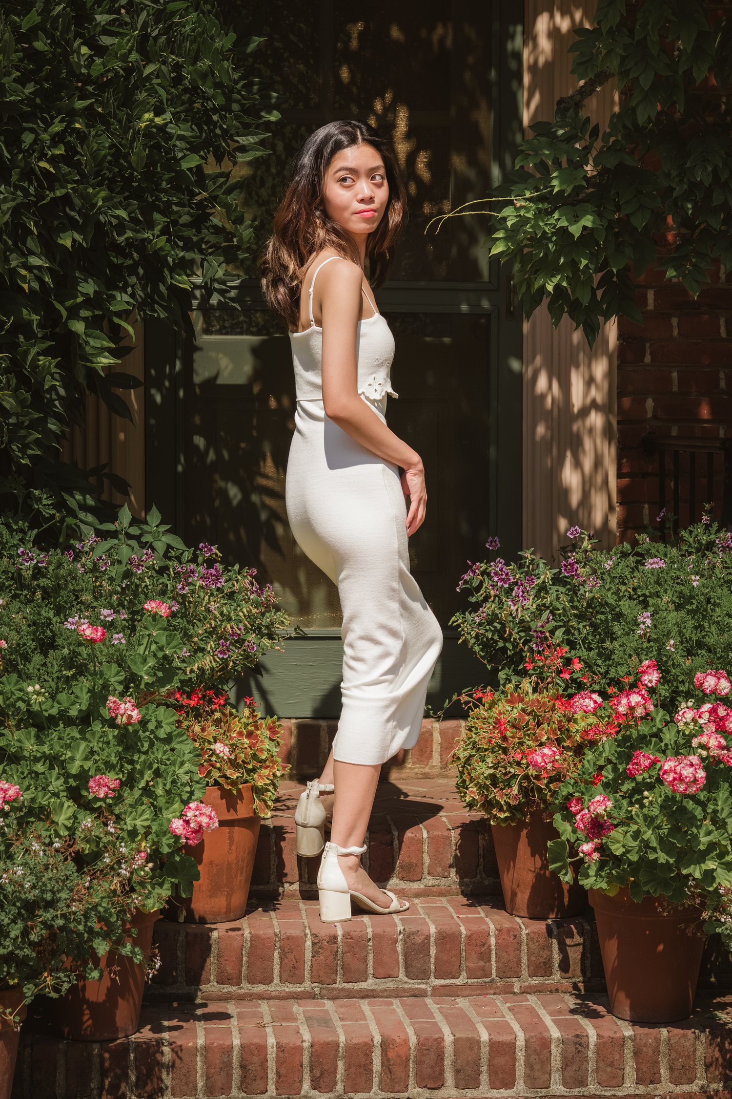 Young woman in white midi dress posing on brick steps surrounded by colorful flower pots and greenery.