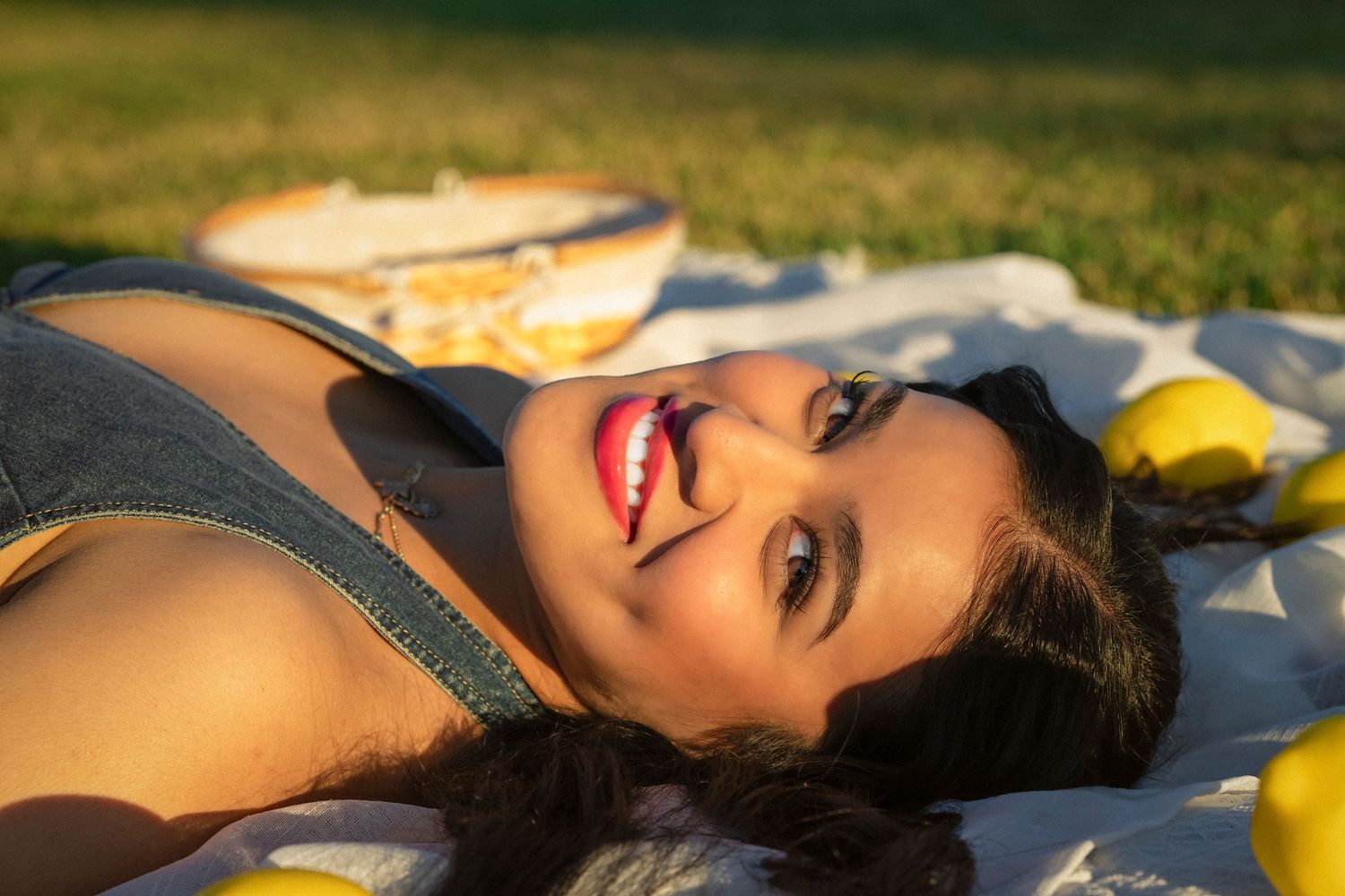 A smiling young woman with red lips lies on a white blanket in a sunny park surrounded by fresh lemons.