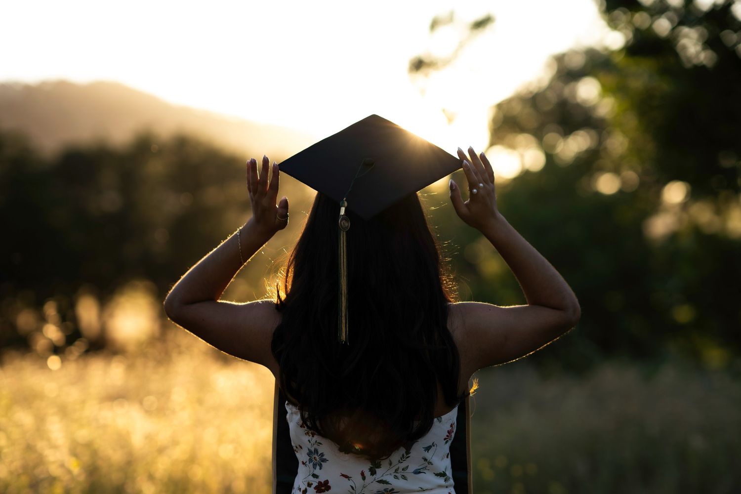 Graduate holding mortarboard cap in sunlit field at golden hour in Brentwood, CA.
