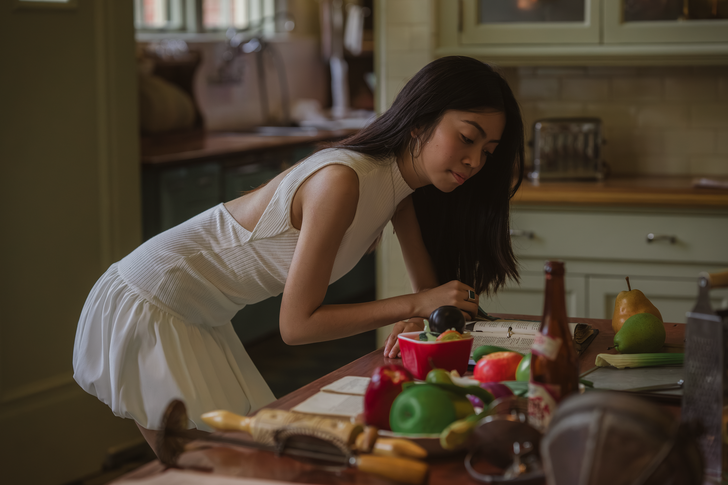 Young Asian woman in white dress leaning over a wooden table with colorful food ingredients in a cozy kitchen.