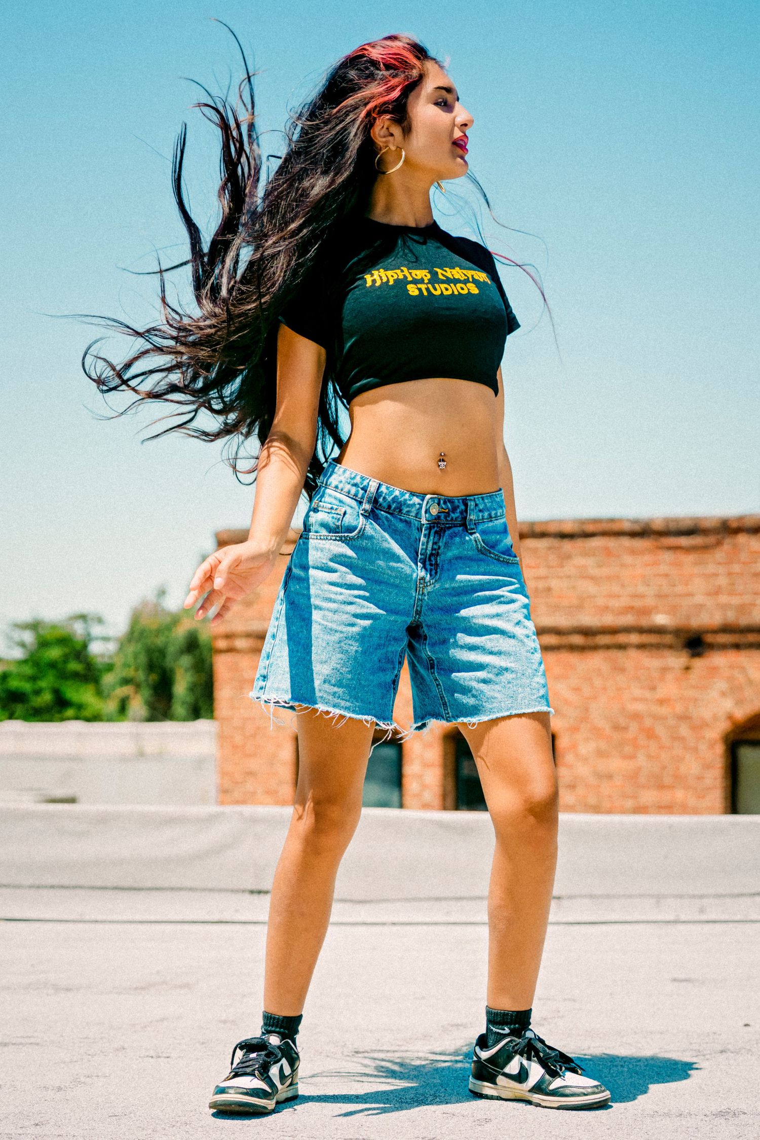 Young woman in black crop top and denim shorts dancing outdoors with hair flowing in the wind on a sunny day.