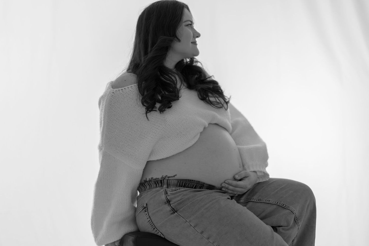 Black and white maternity photo of pregnant woman sitting and cradling her baby bump.